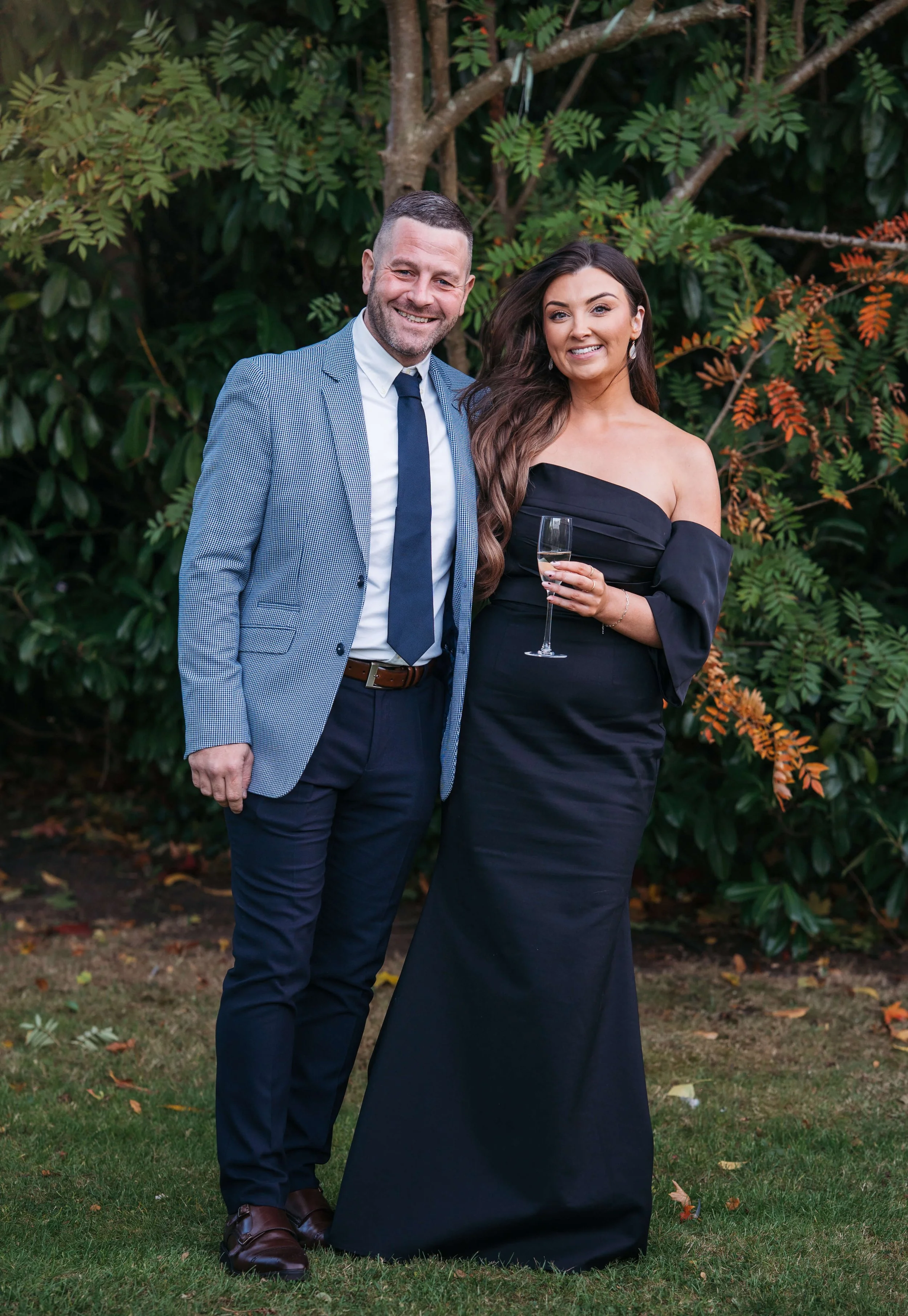 A man and woman in formal attire standing outdoors in front of green foliage, smiling at the camera. The woman is holding a glass of champagne.