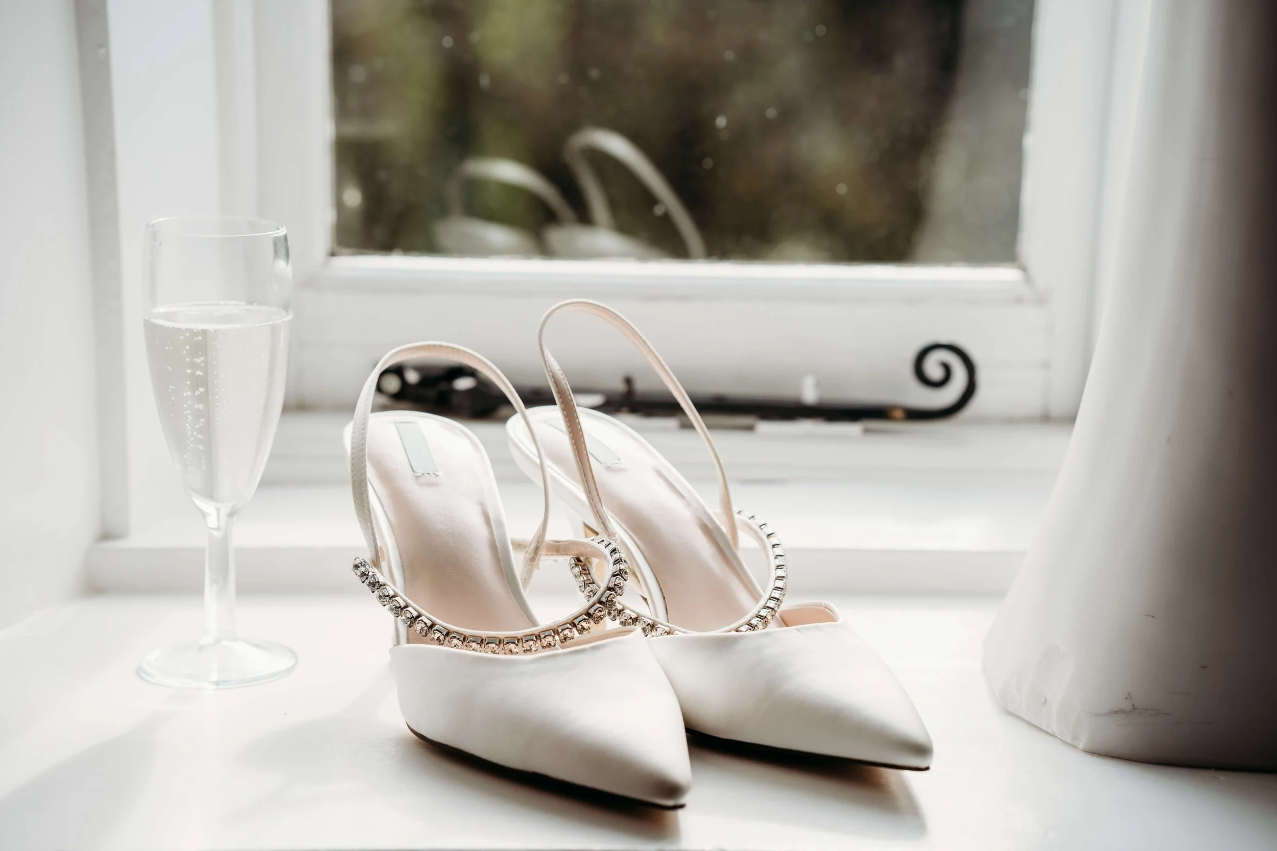 A pair of white high-heeled shoes with rhinestone embellishments, a glass of champagne, and white curtains by a window.