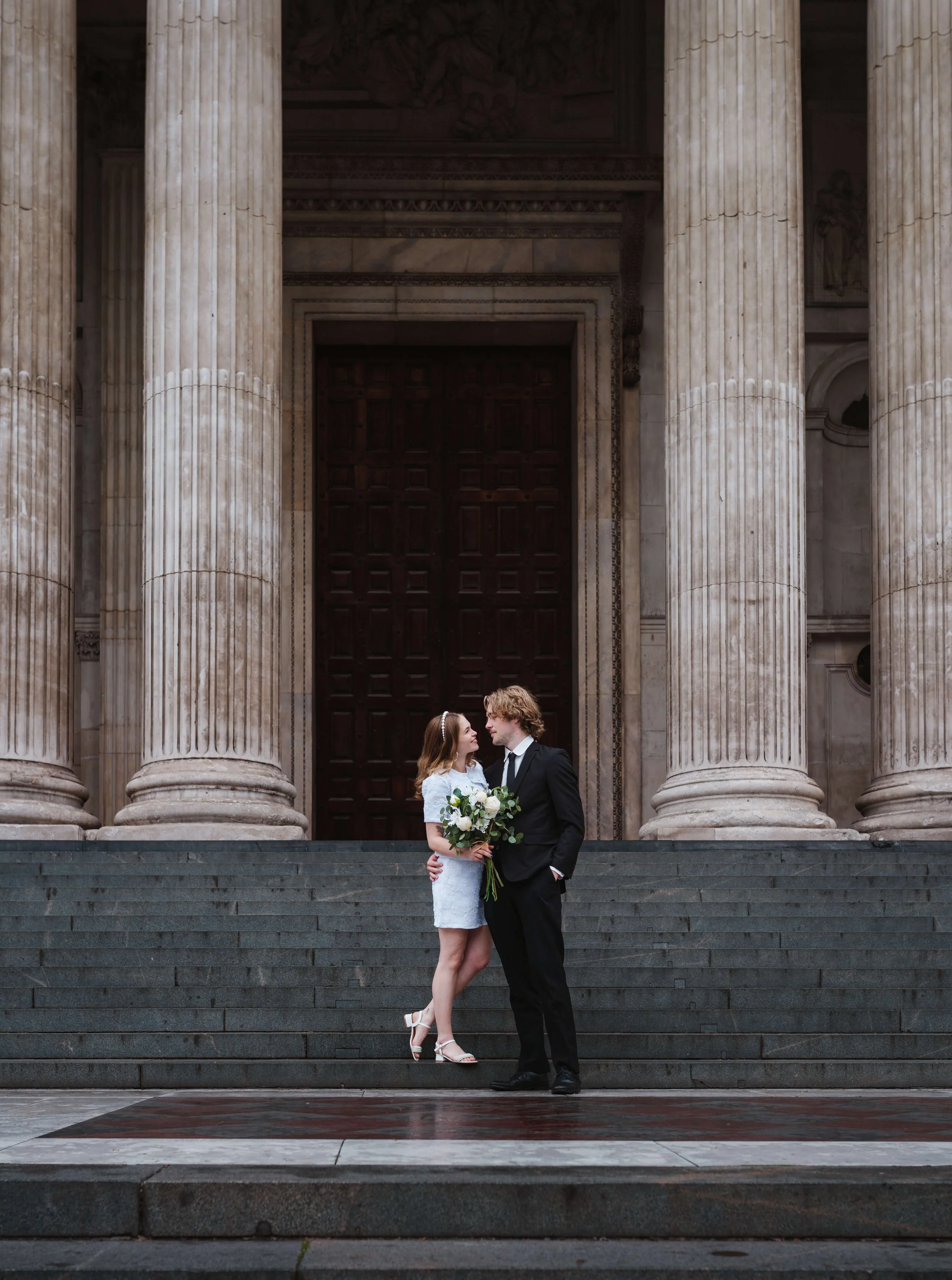 A bride in a white dress holding a bouquet and a groom in a black suit sharing a moment on the stairs outside a building with large stone columns.