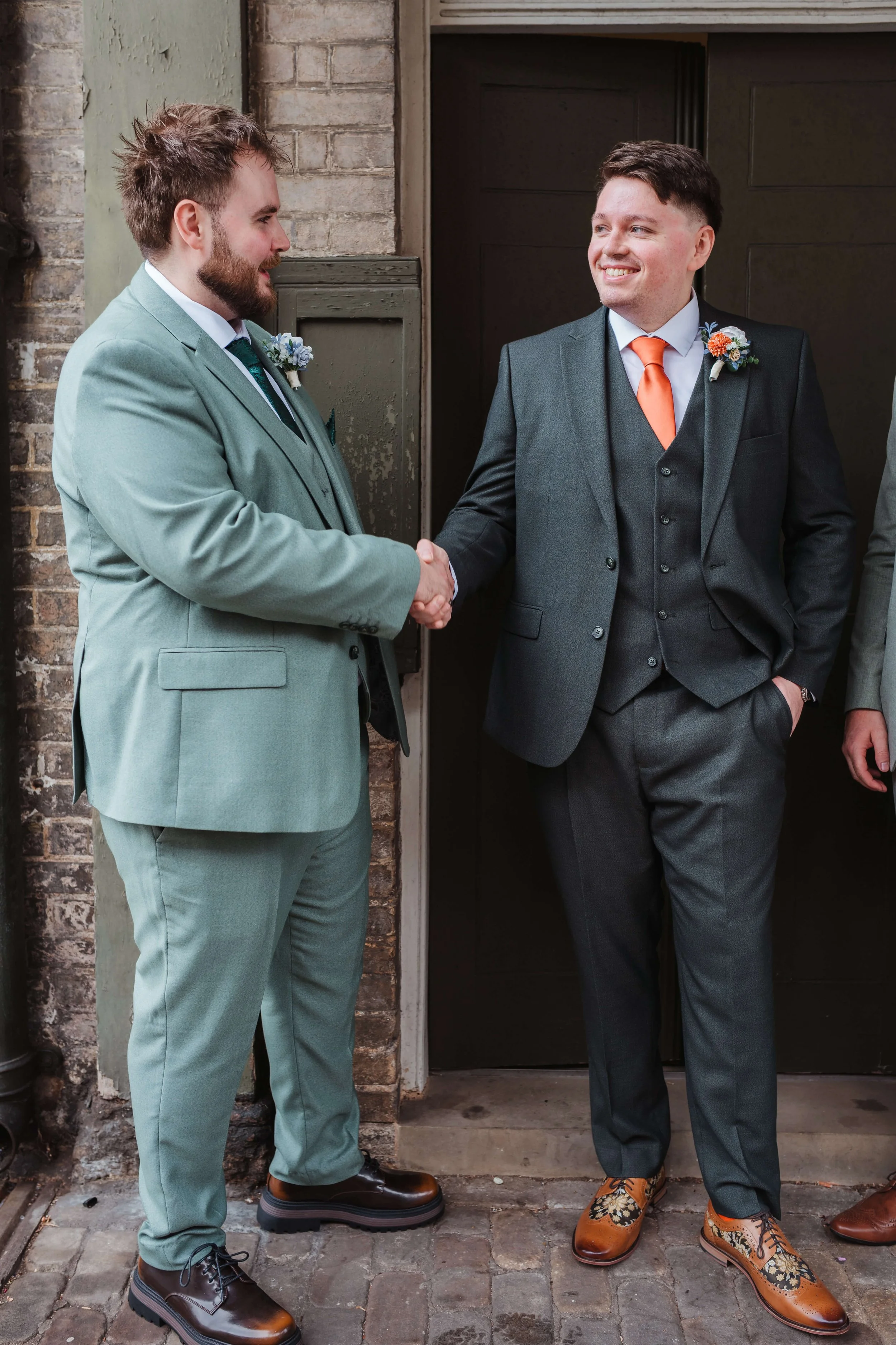 Two men in suits shaking hands outside a brick building, smiling at each other, with boutonnières on their lapels.