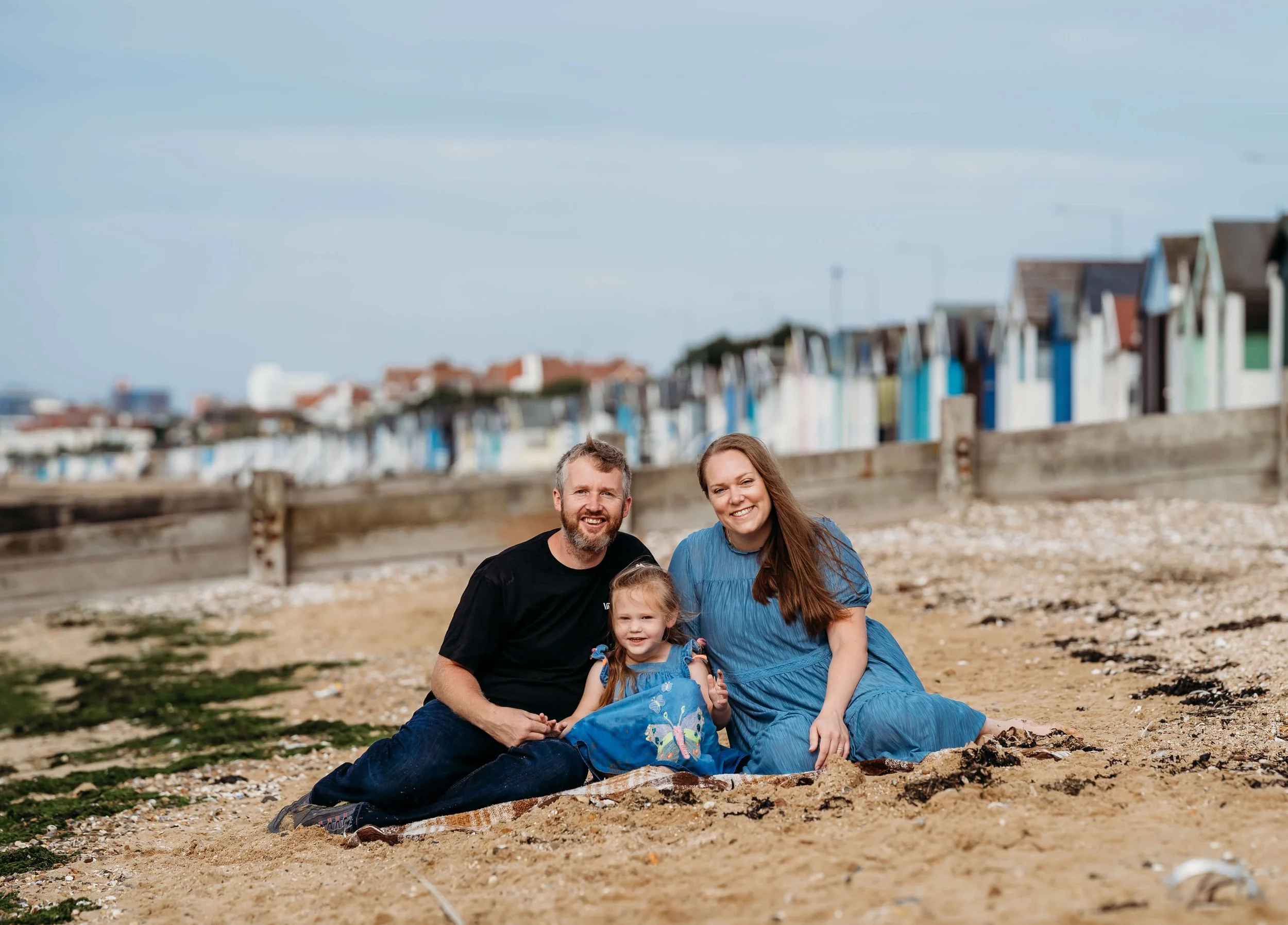 A family of three sitting on the beach, smiling at the camera with colorful houses in the background.