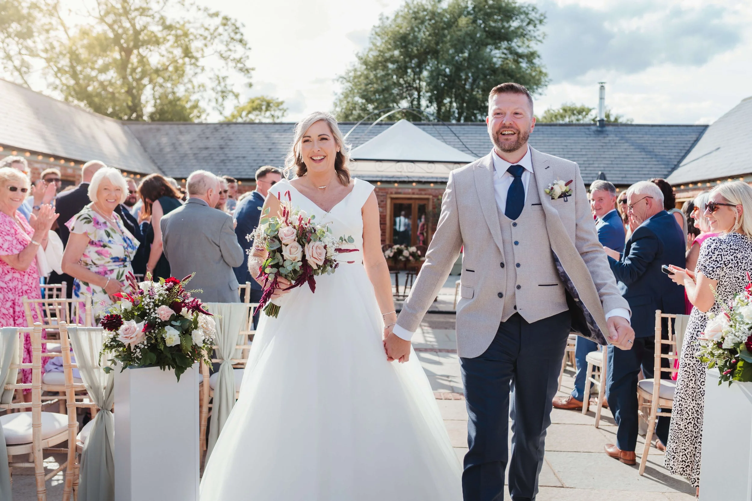 A bride and groom walking hand in hand after their wedding ceremony outdoors, surrounded by smiling guests, with flowers and chairs along the aisle.