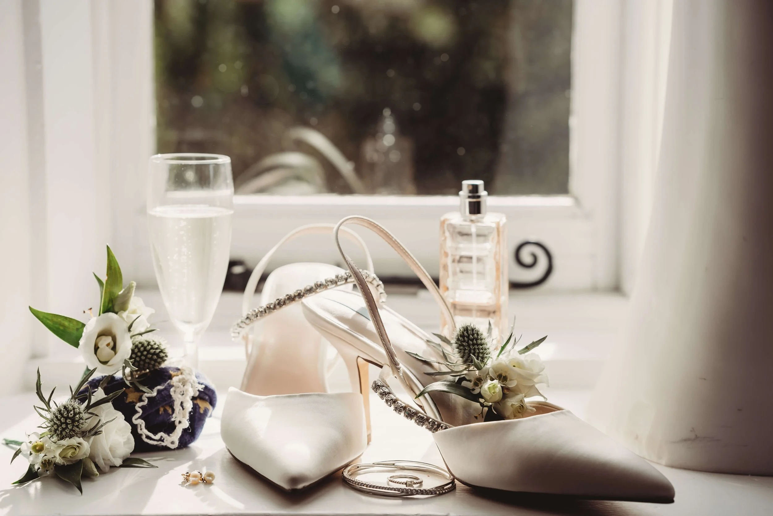 Wedding shoes, flowers, jewelry, perfume, and a glass of champagne on a windowsill.