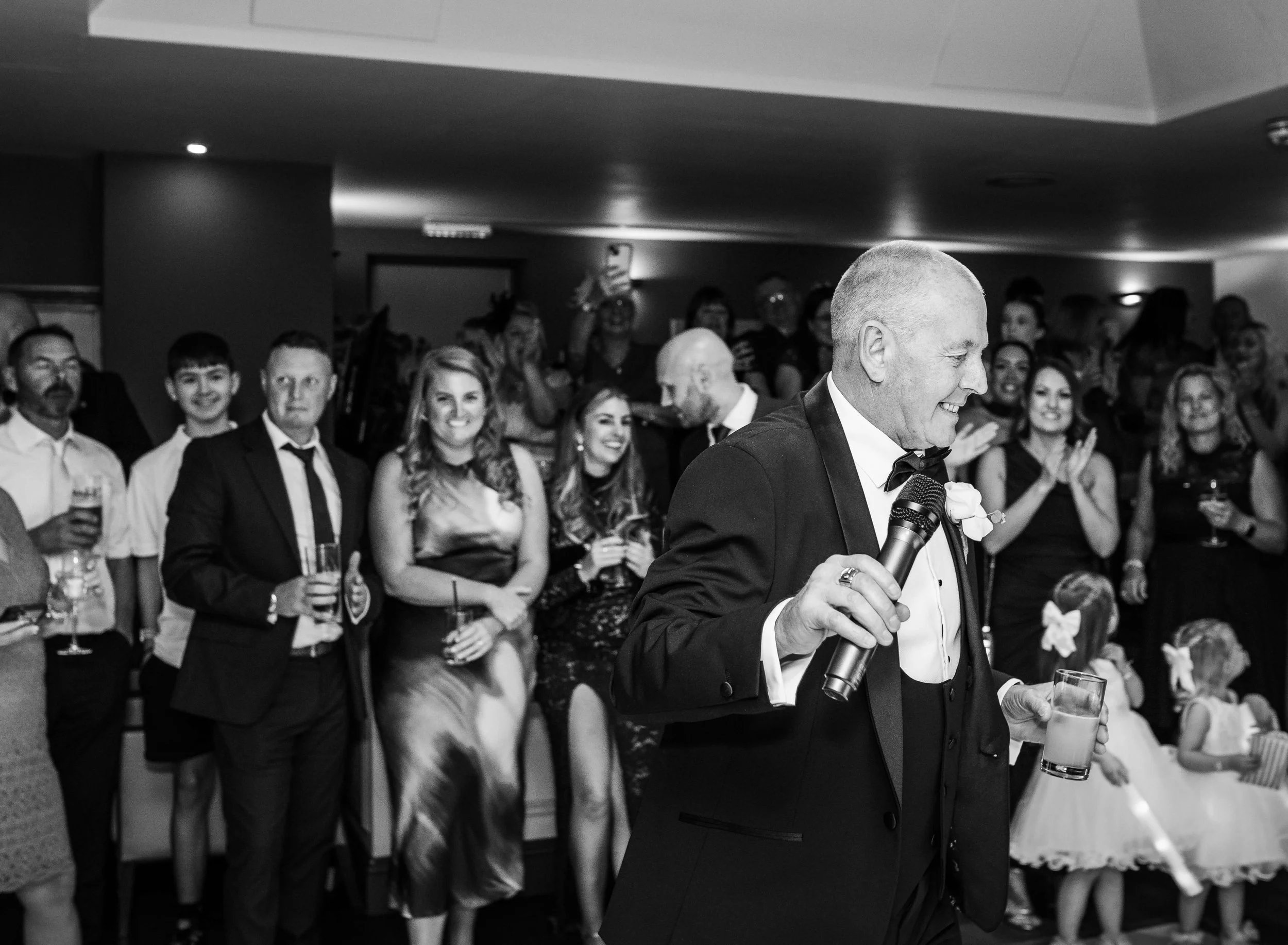 A man in a tuxedo holding a microphone and a glass, smiling as he gives a speech, surrounded by an audience clapping and smiling at a formal celebration or wedding reception.