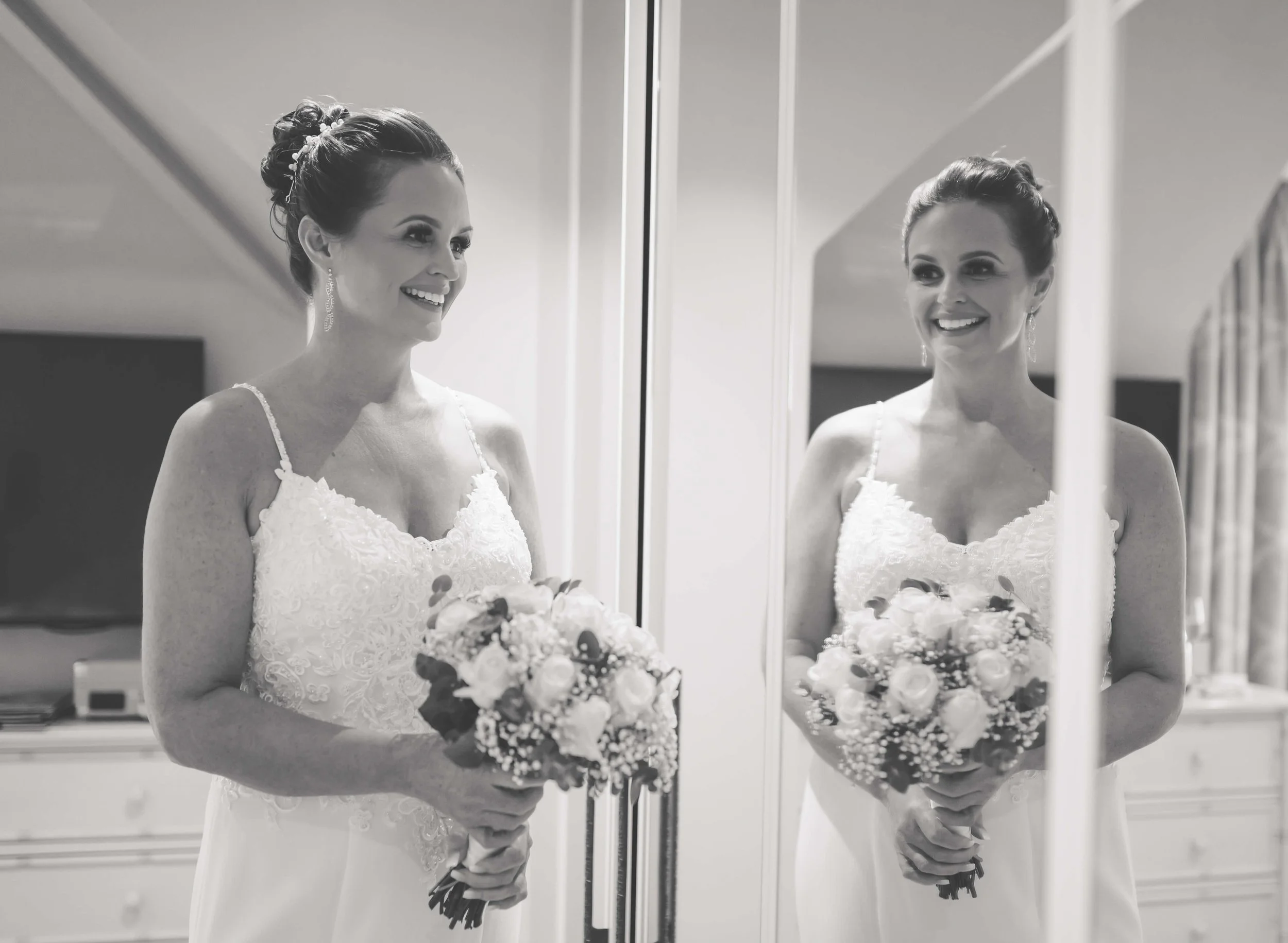 A woman in a white wedding dress holding a bouquet, standing in front of a mirror, smiling at her reflection.