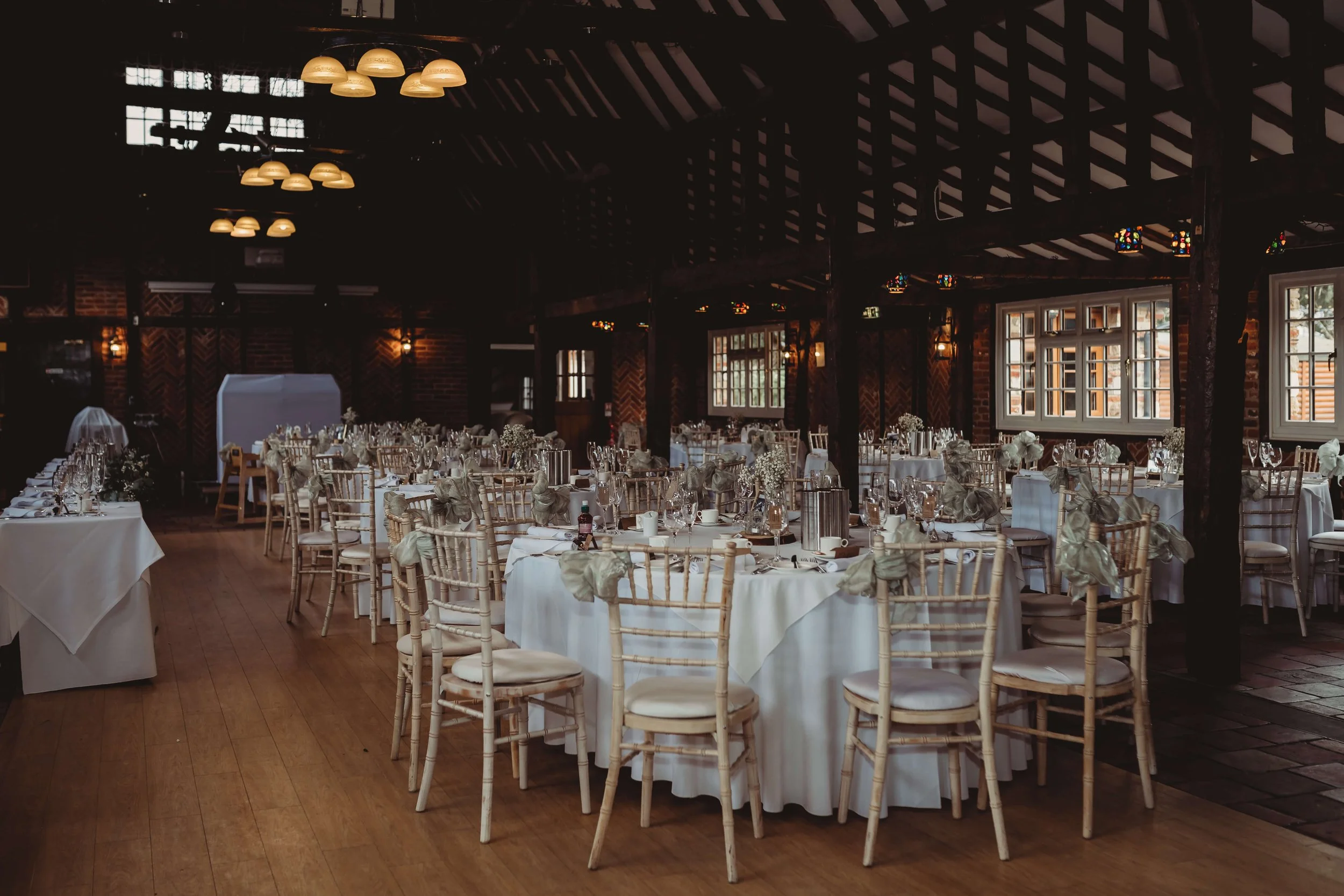 Wedding reception hall with round tables covered in white tablecloths, decorated with floral centerpieces and set with wine glasses, plates, and silverware. Wooden chairs with white cushions surround the tables. The room has dark brick walls, large w