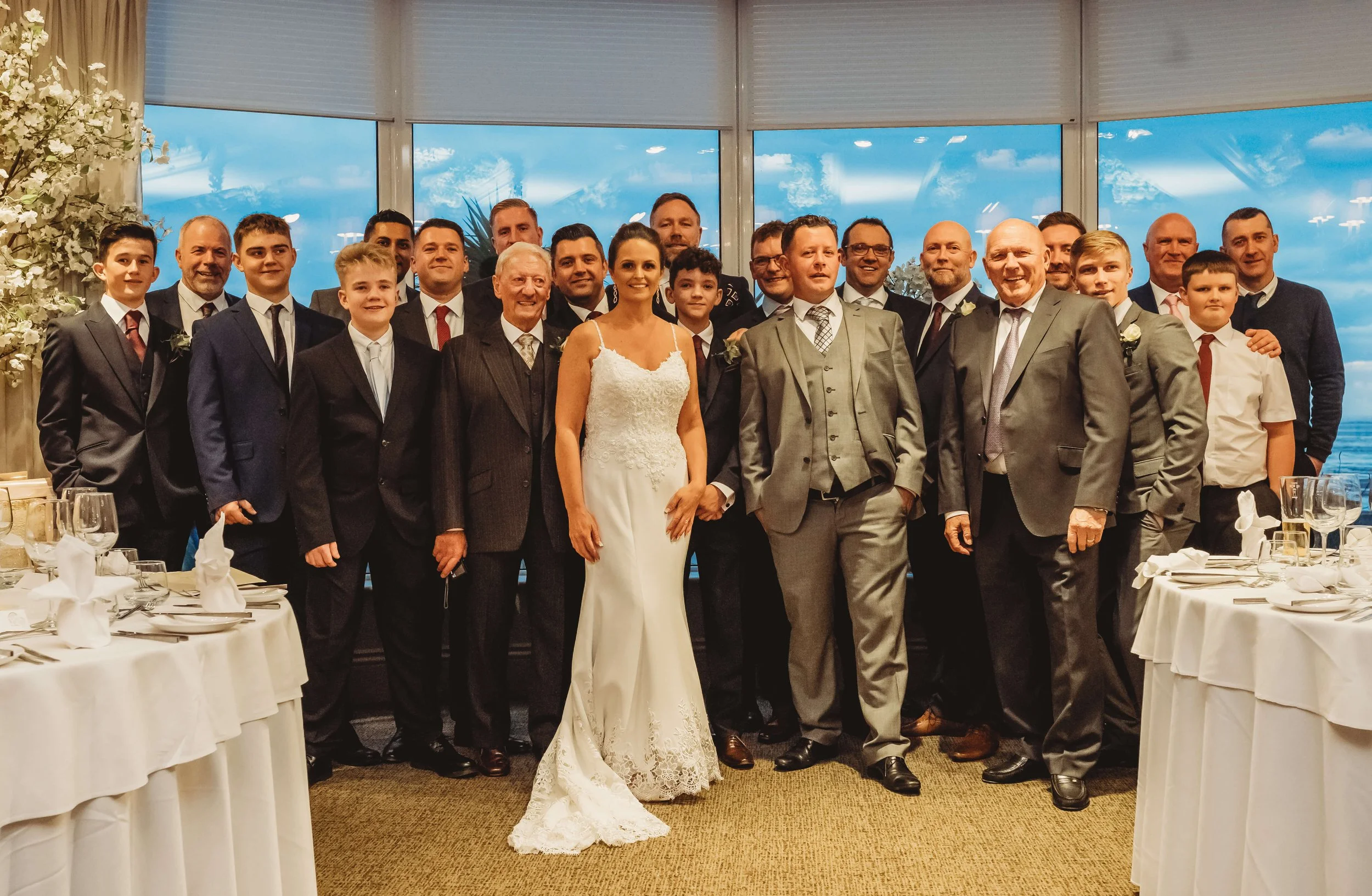 Group of people at a wedding reception, including a bride in a white wedding dress and a groom in a gray suit, indoors with a large window and a cloudy sky outside.