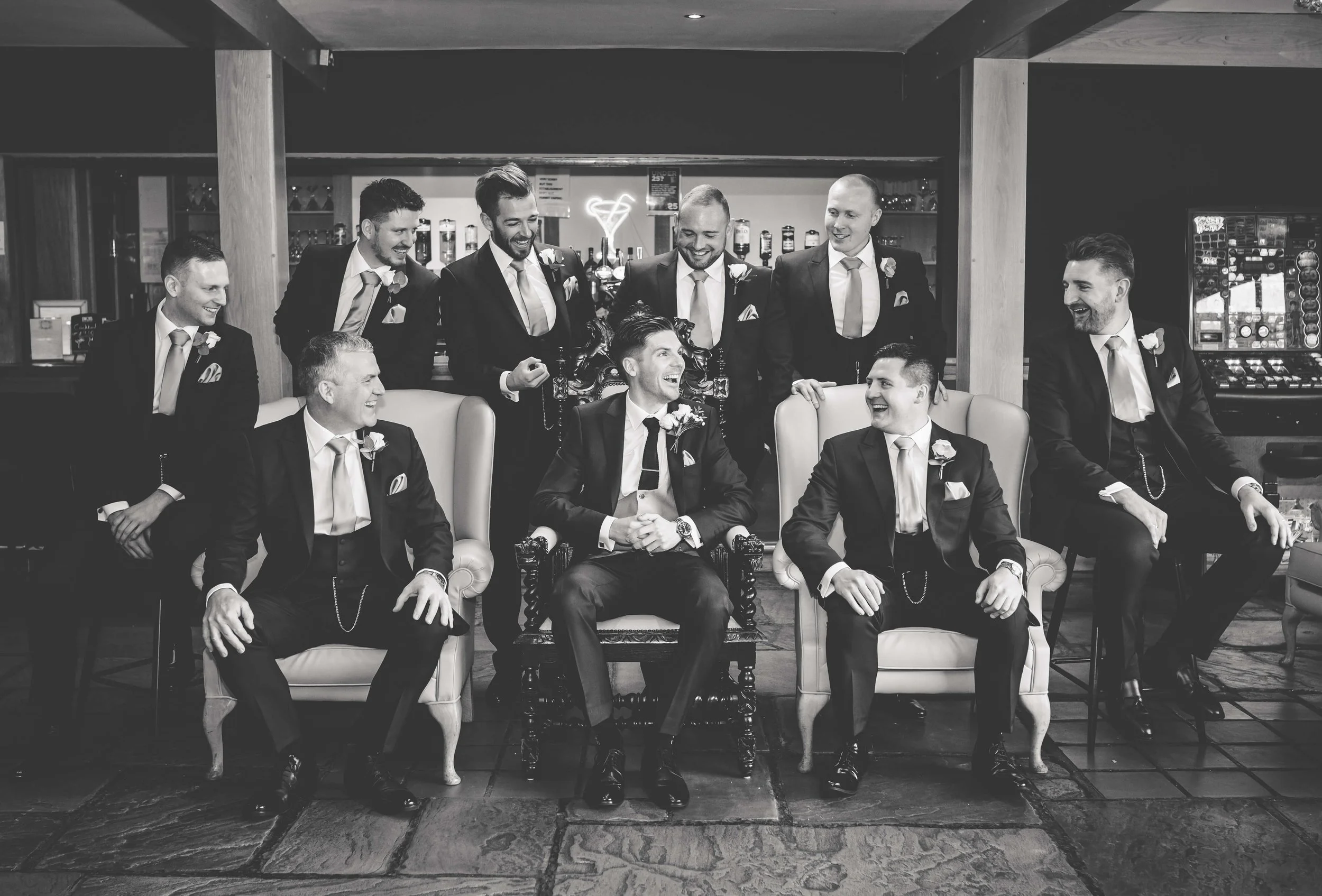 Black and white photo of a group of nine men, dressed in suits with boutonnieres, gathered in a lounge or bar area. Four men are seated on sofas, with the one in the center and to his right smiling and looking at each other, while five men stand behi
