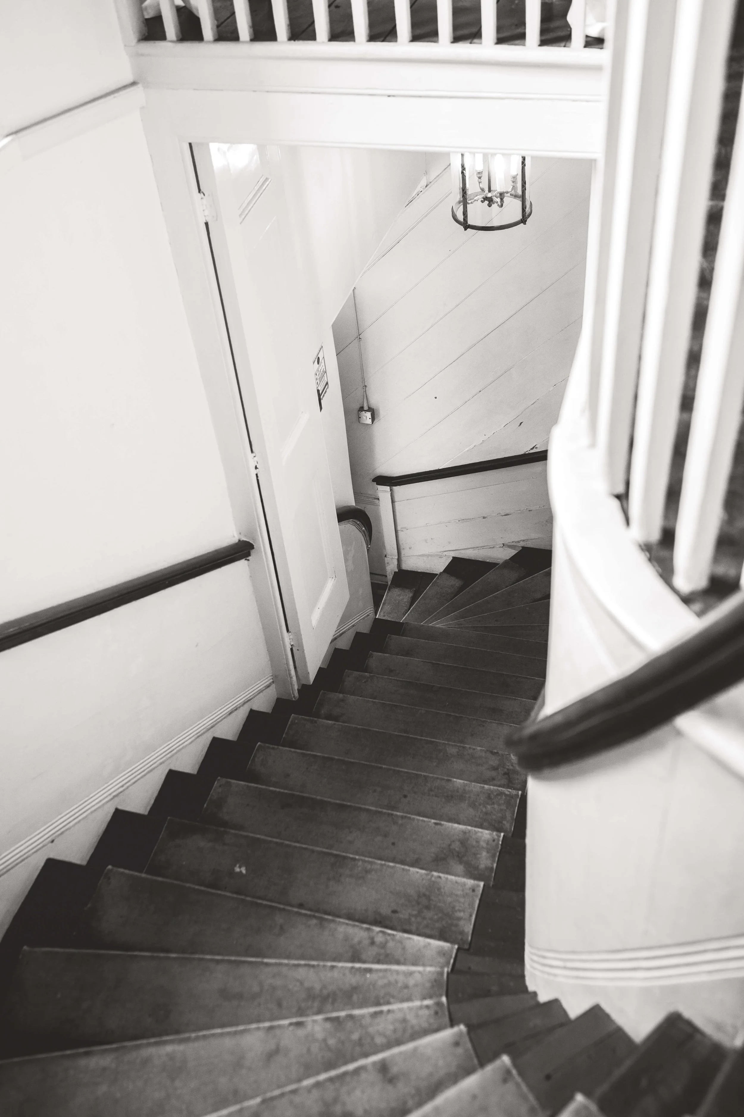 Black and white photo of a spiral staircase viewed from the top, with wooden steps and white railing, leading down to a door at the bottom.