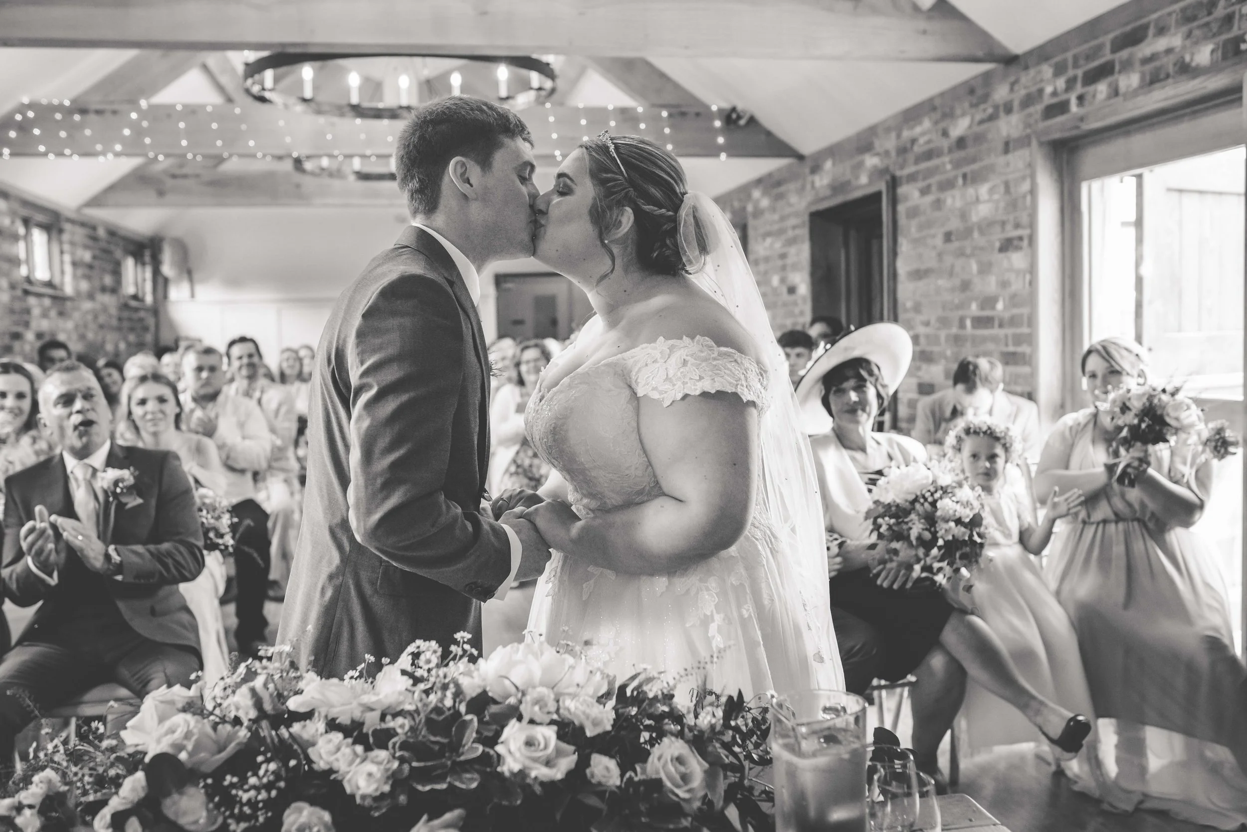 A bride and groom kiss during their wedding ceremony in a rustic venue with brick walls, while wedding guests look on in the background.
