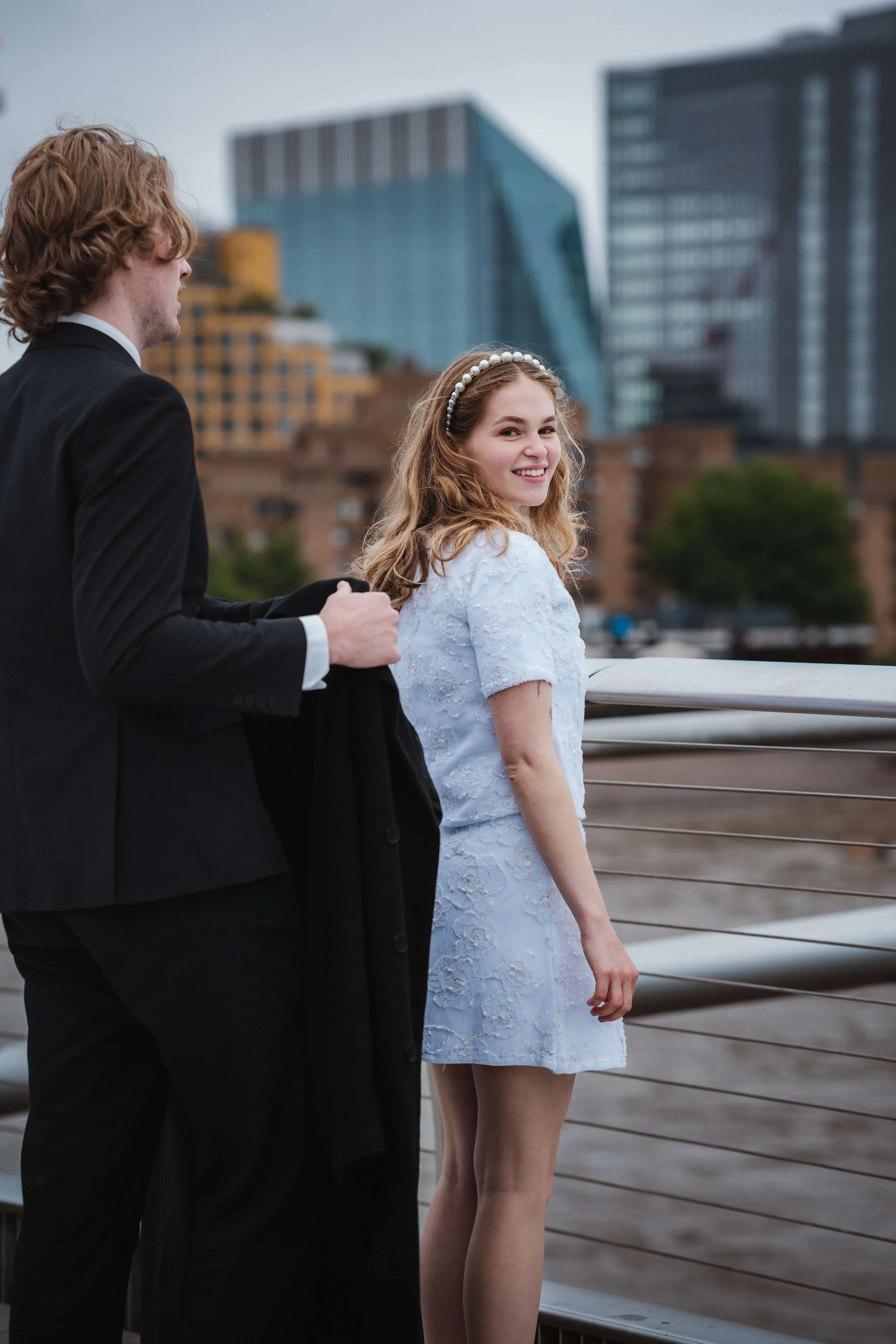 A young woman in a white dress smiling and looking back at a man in a black suit on a city balcony with tall modern buildings in the background.