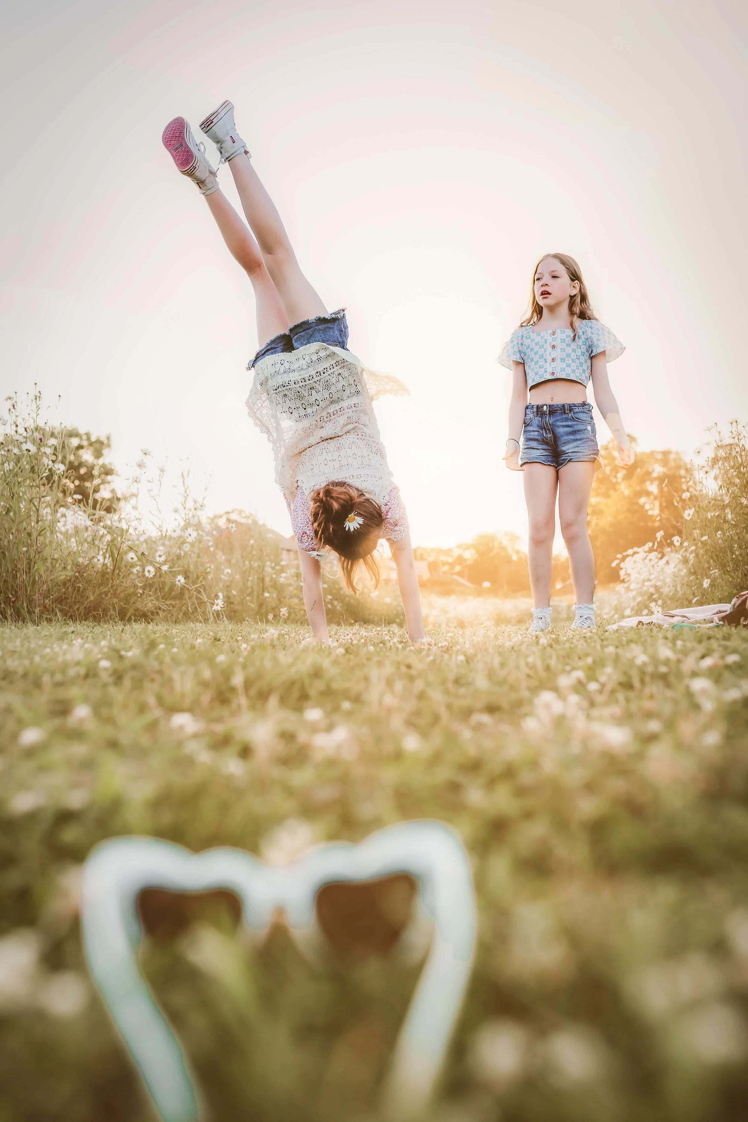 Two young girls playing outdoors in a field at sunset, with one girl performing a handstand and the other girl standing nearby. A pair of sunglasses is visible in the foreground.