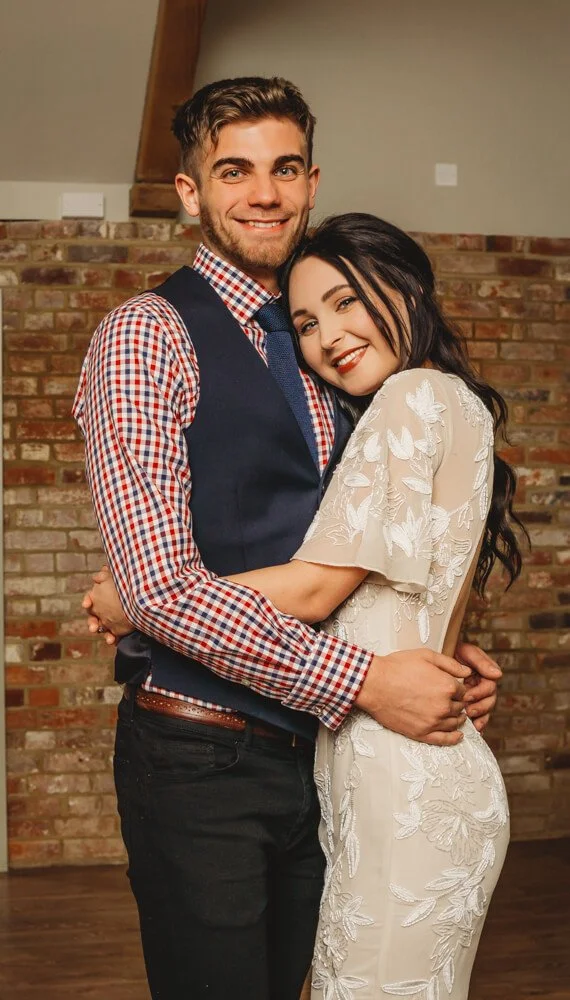 A smiling couple hugging each other indoors, with a brick wall background. The man has short brown hair and is wearing a checkered shirt with a vest, while the woman has long dark hair and is wearing a cream-colored dress with floral embroidery.