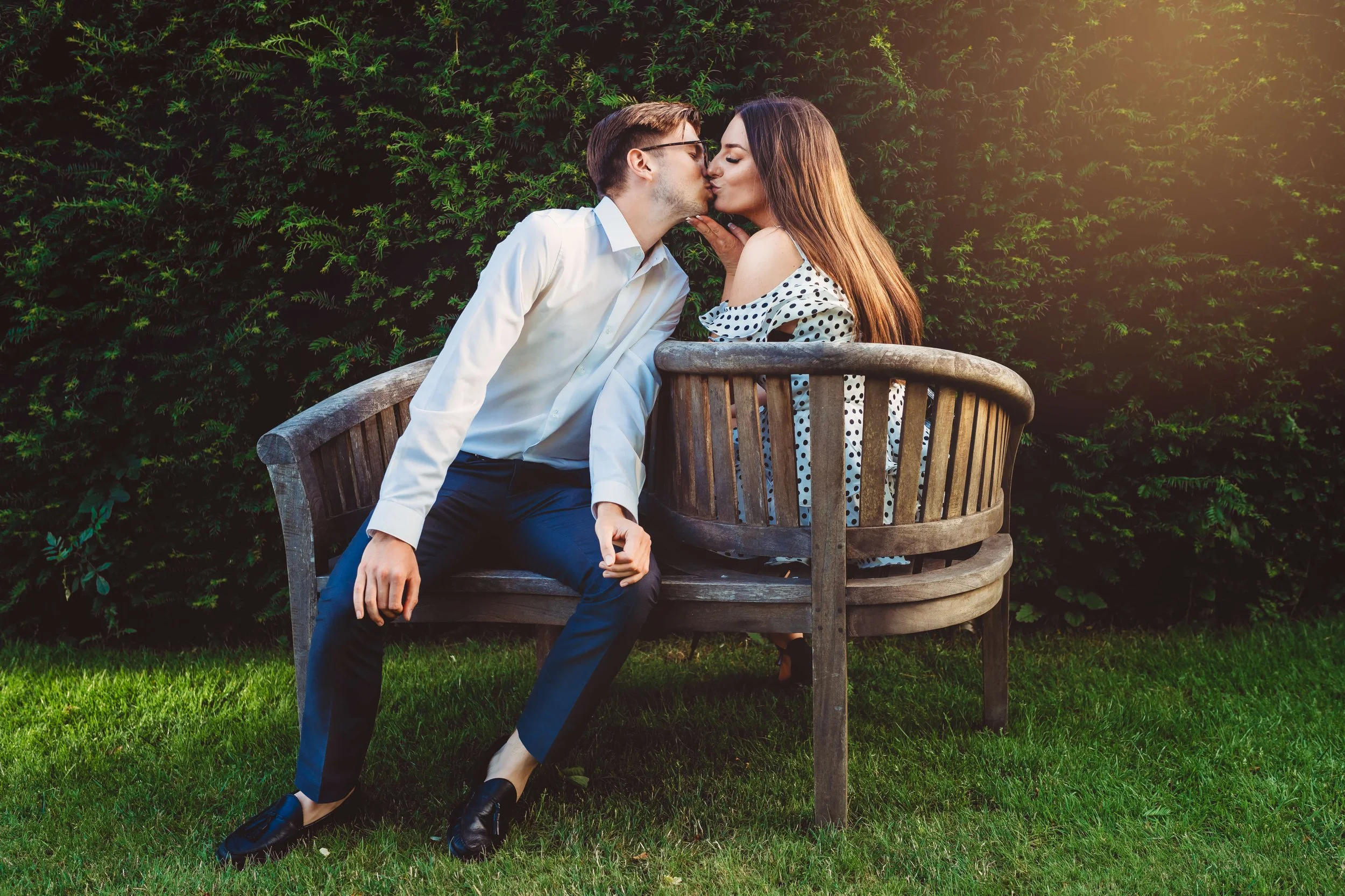 A man and a woman share a kiss on a park bench, with green bushes in the background and sunlight on the right side.