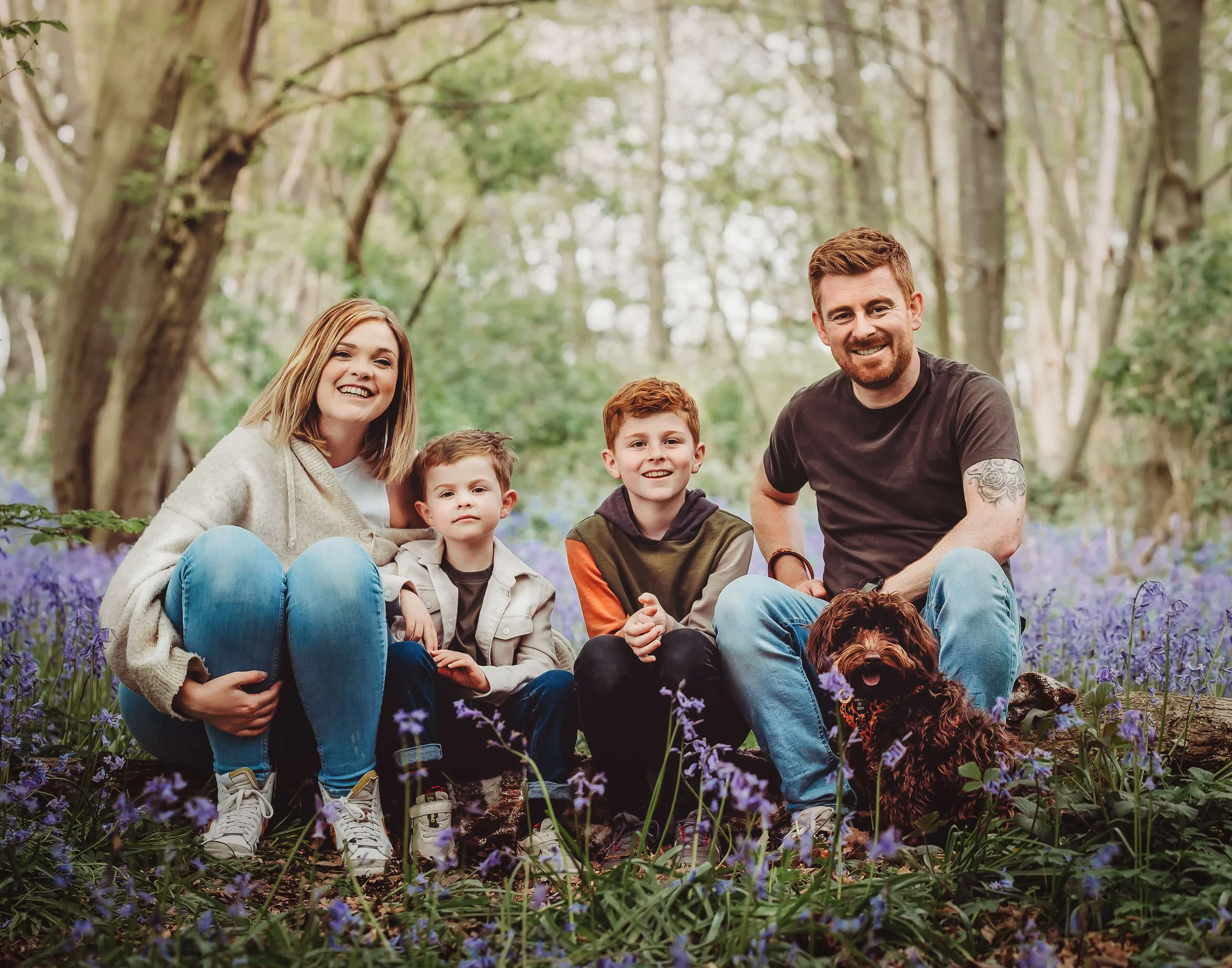 A family of four with two young boys and their parents, along with a dog, sitting outdoors among purple flowers and trees in a forest.