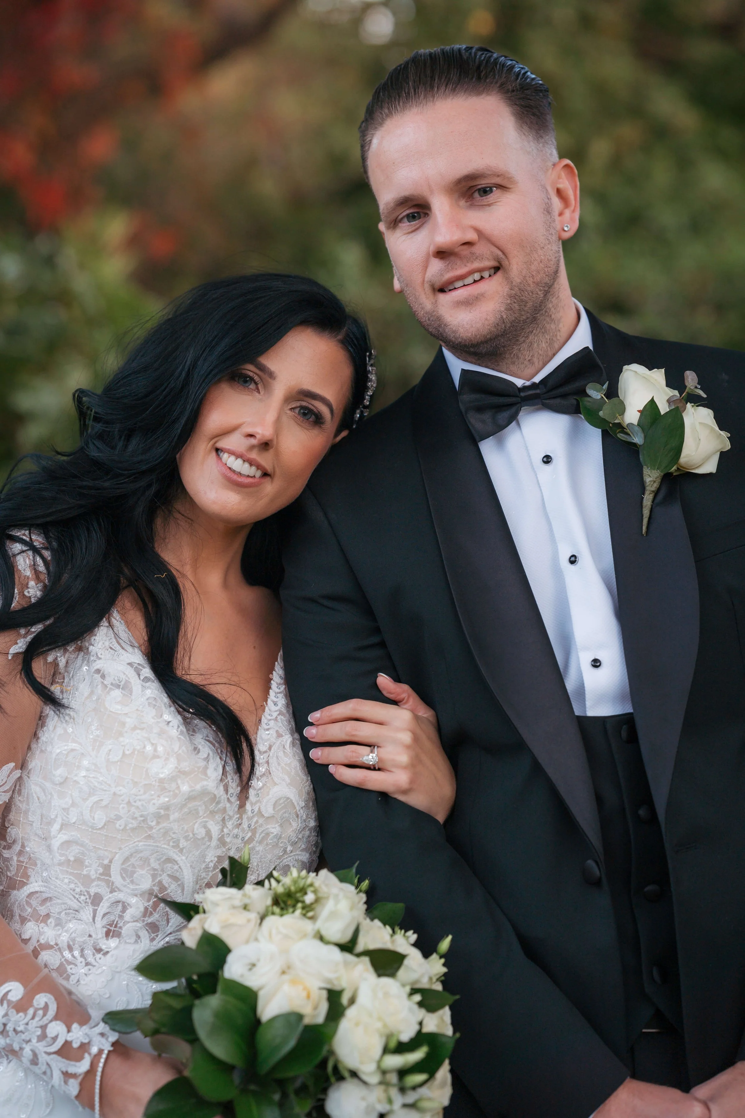 A bride and groom pose together outdoors on their wedding day, with the bride holding a bouquet of white roses and the groom wearing a black tuxedo with a white shirt and black bow tie. The bride has long black hair and the groom has short slicked-ba