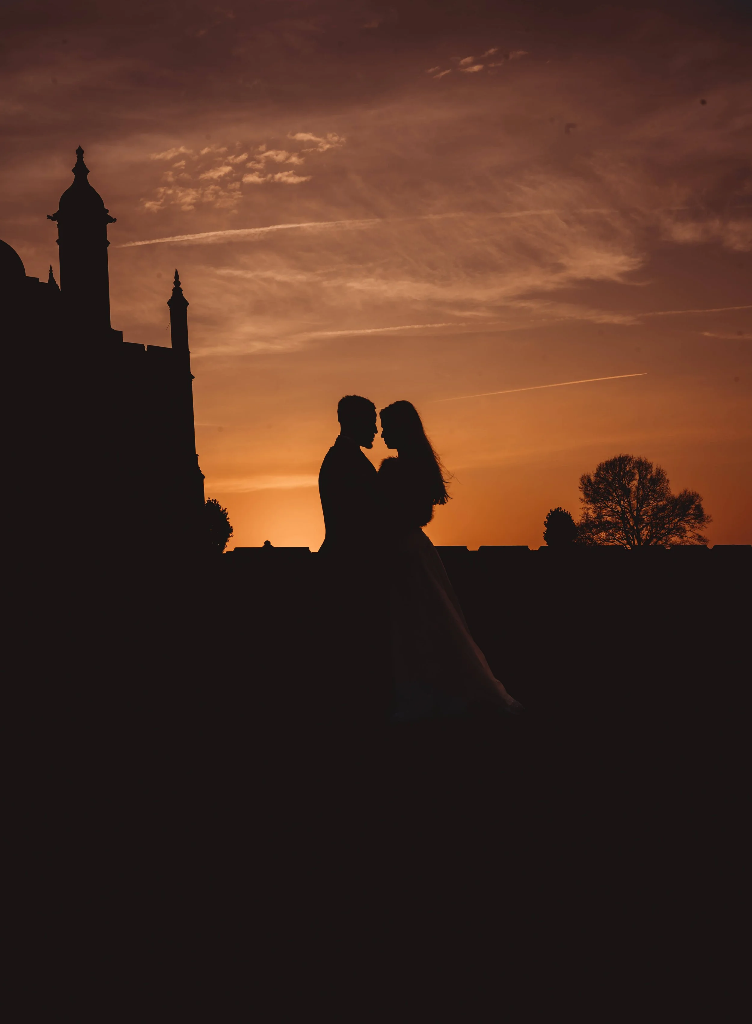 Silhouette of a couple standing close together during sunset, with a historic building and trees in the background.