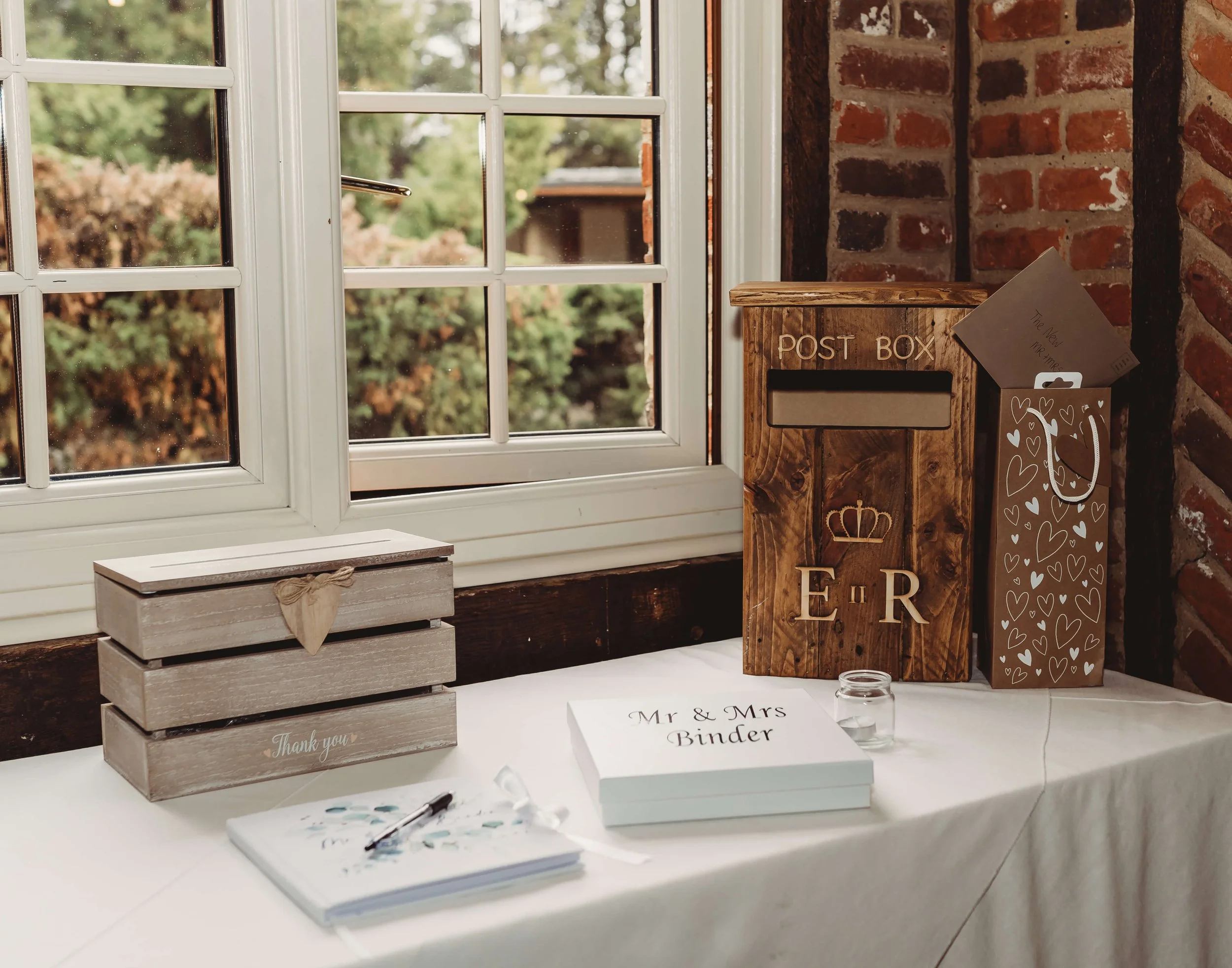 A wooden mailbox labeled 'Post Box' and two cardboard boxes with hearts and crown symbols are placed on a table near a window, along with a binder for Mr. and Mrs., a mason jar, a small candle, a notebook, and a pen.