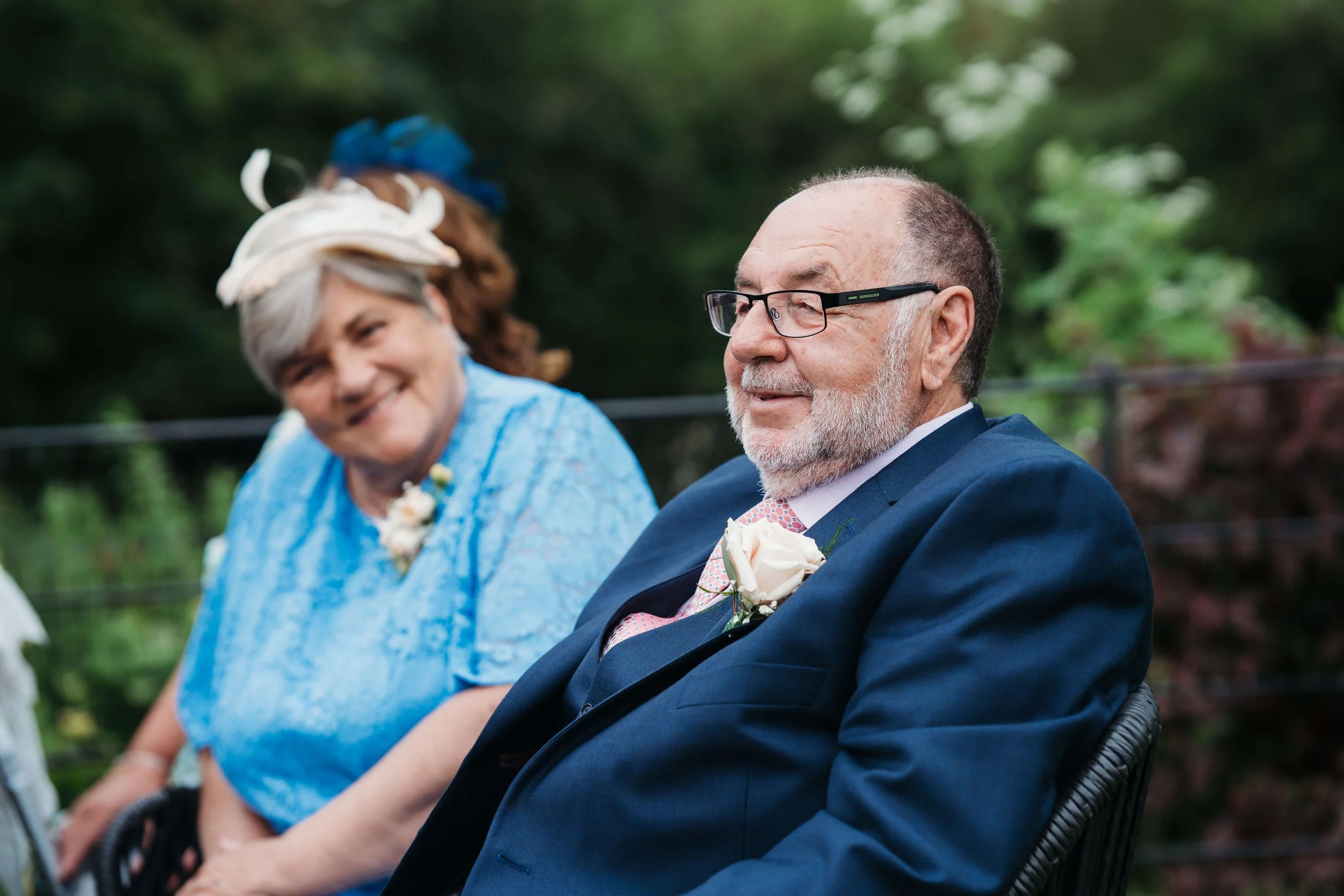 An elderly man wearing a suit and glasses sitting outdoors, with an elderly woman in a blue dress next to him, both smiling at a social event, with greenery in the background.