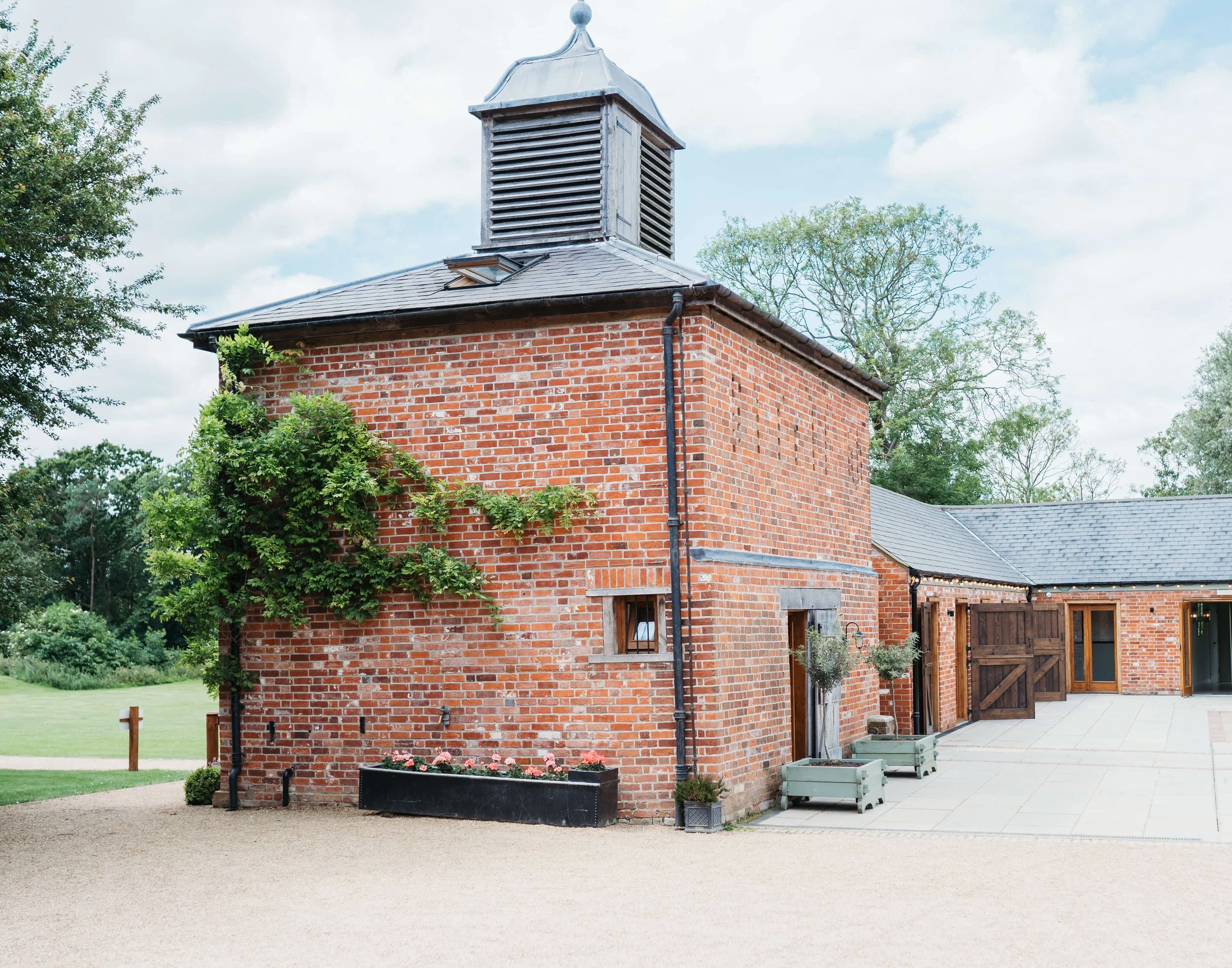 A red brick building with a small window, a door, and plants outside, surrounded by greenery under a cloudy sky.