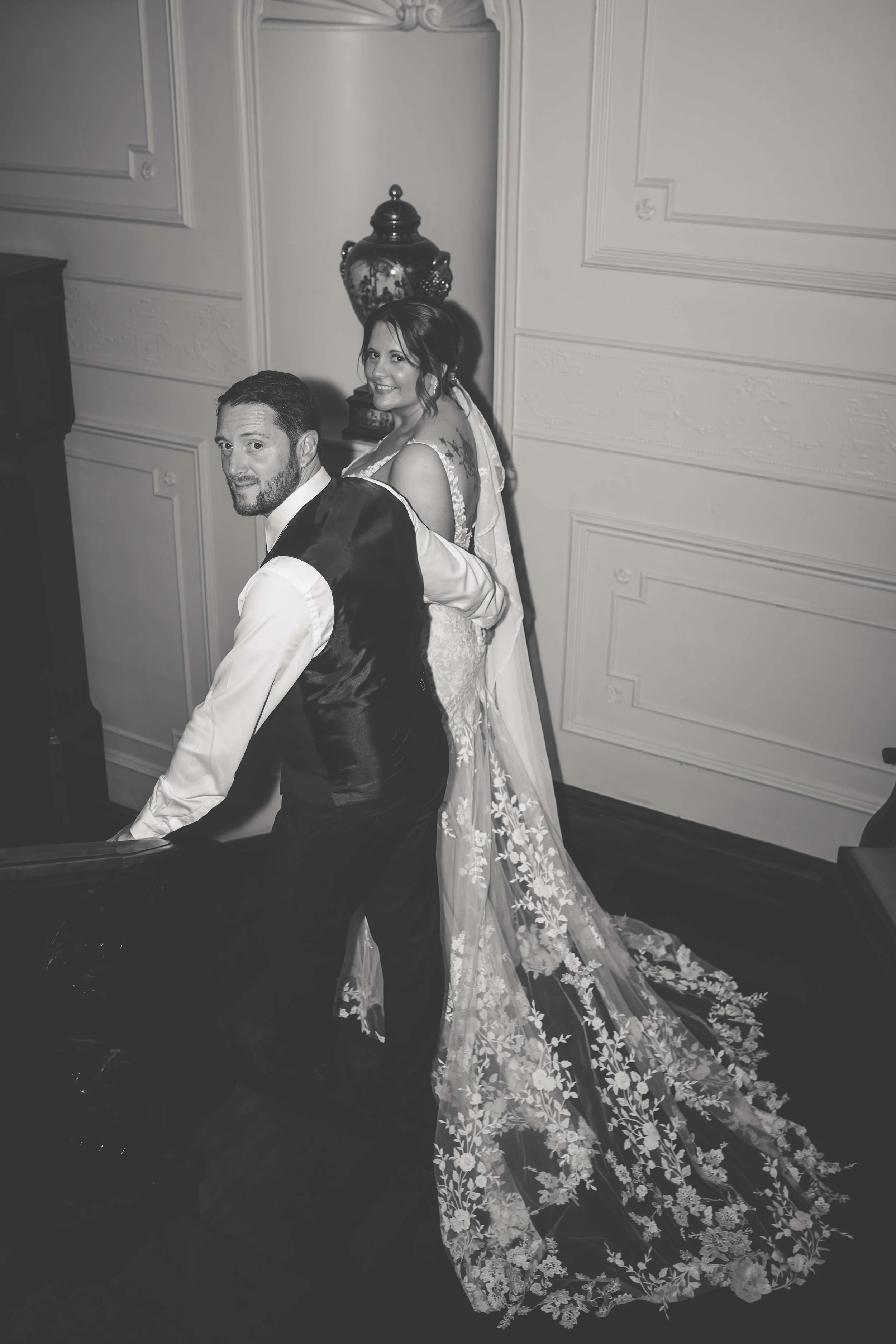Black-and-white photo of a bride and groom, where the groom is leaning on a stair banister and the bride stands behind him, smiling, in an elegantly decorated room.