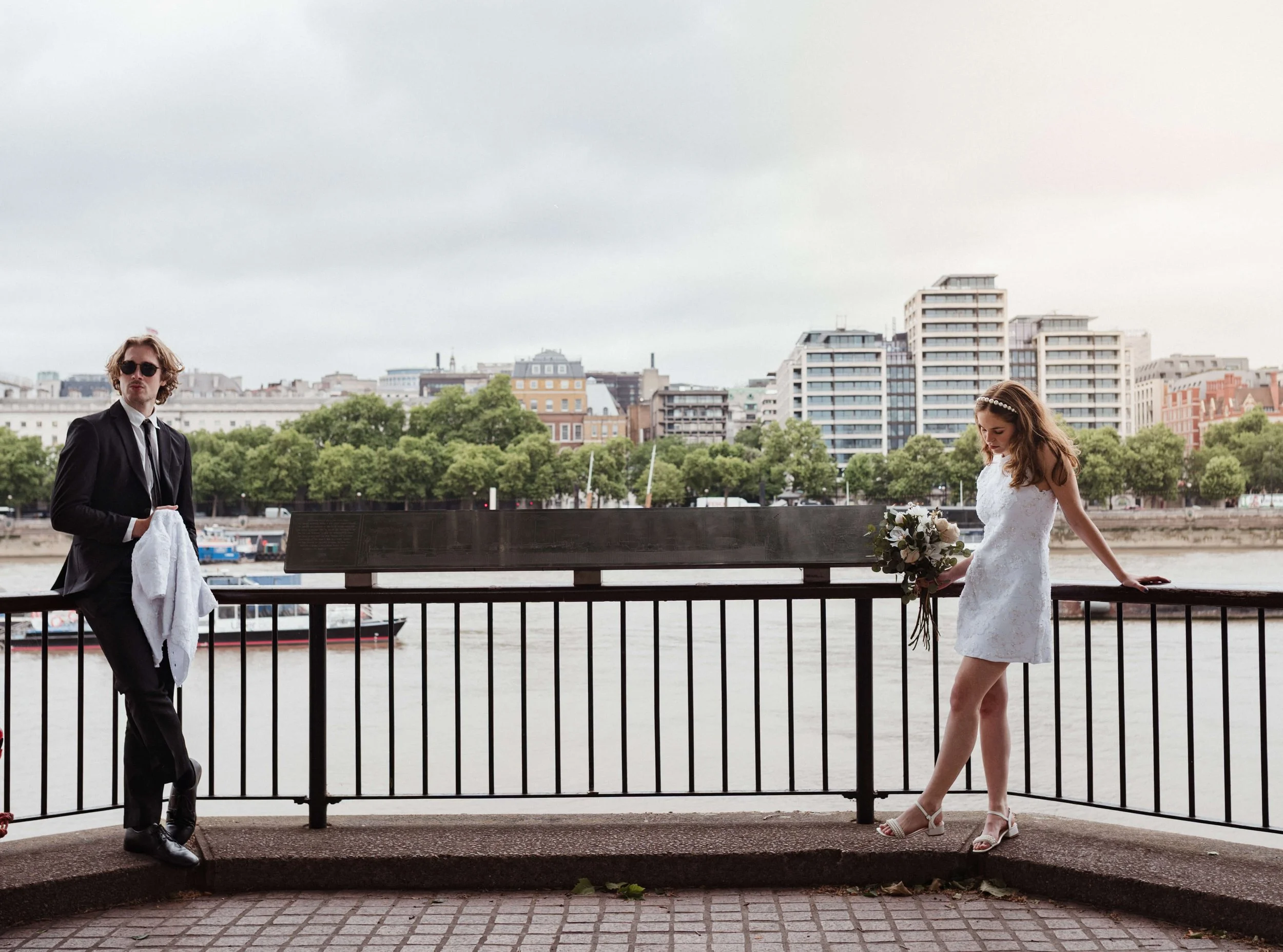 A couple stands by the river on a cloudy day, with the woman in a white dress holding a bouquet and the man in a black suit holding a white cloth, with modern buildings and green trees in the background.