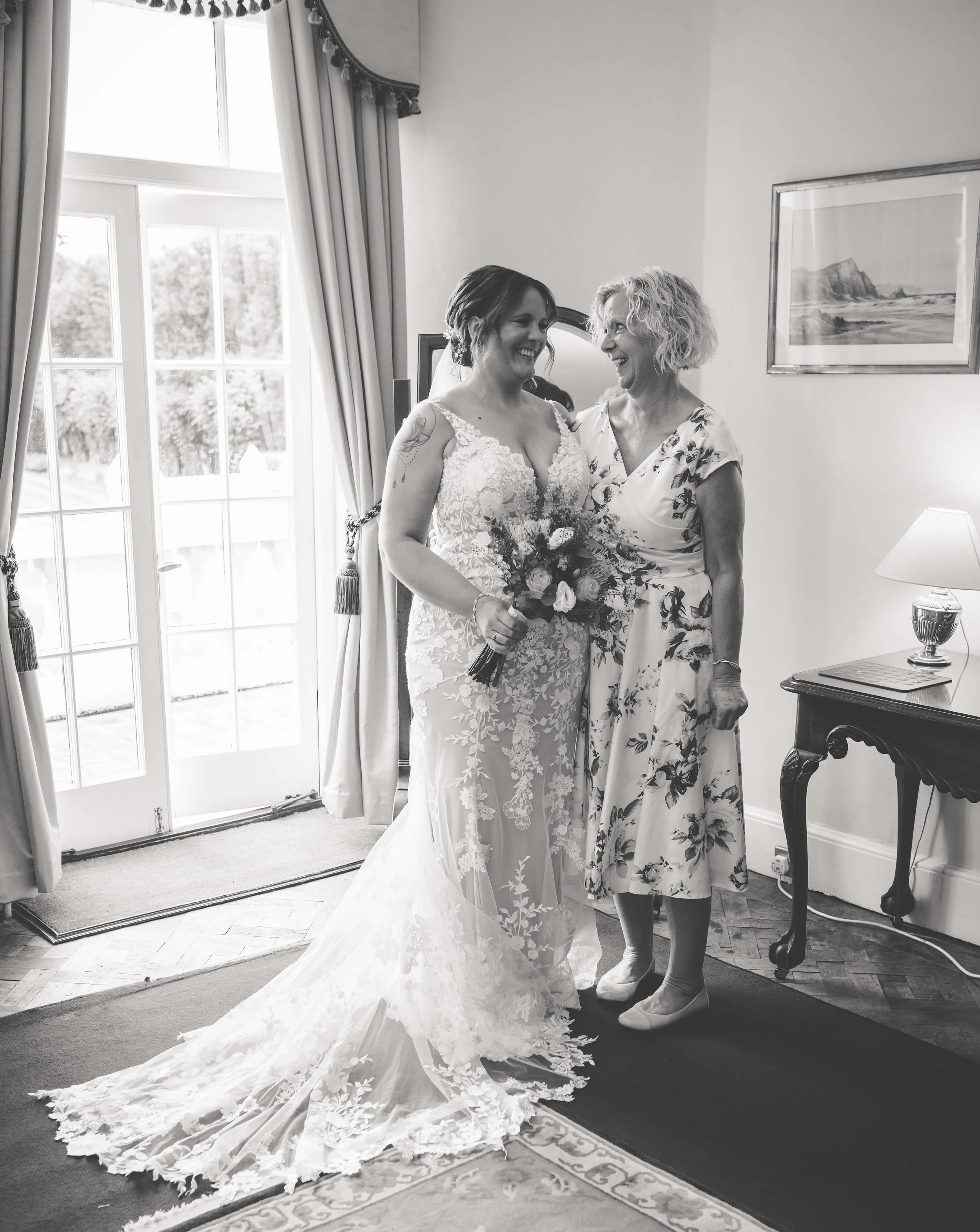 Black and white photo of a bride in a lace wedding gown holding a bouquet, smiling and sharing a joyful moment with an older woman in a floral dress, inside a room with a large window, curtains, and a framed picture on the wall.
