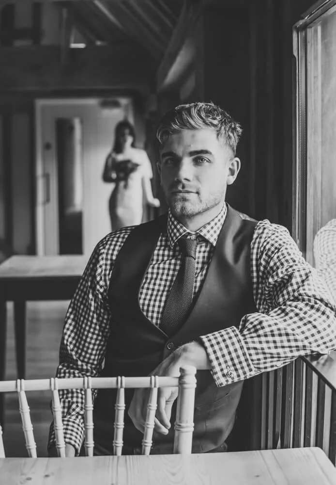 A man with short hair, dressed in a checkered shirt and vest, sitting at a table by a window, looking at the camera, with a woman in a blurred background inside a rustic-style room.