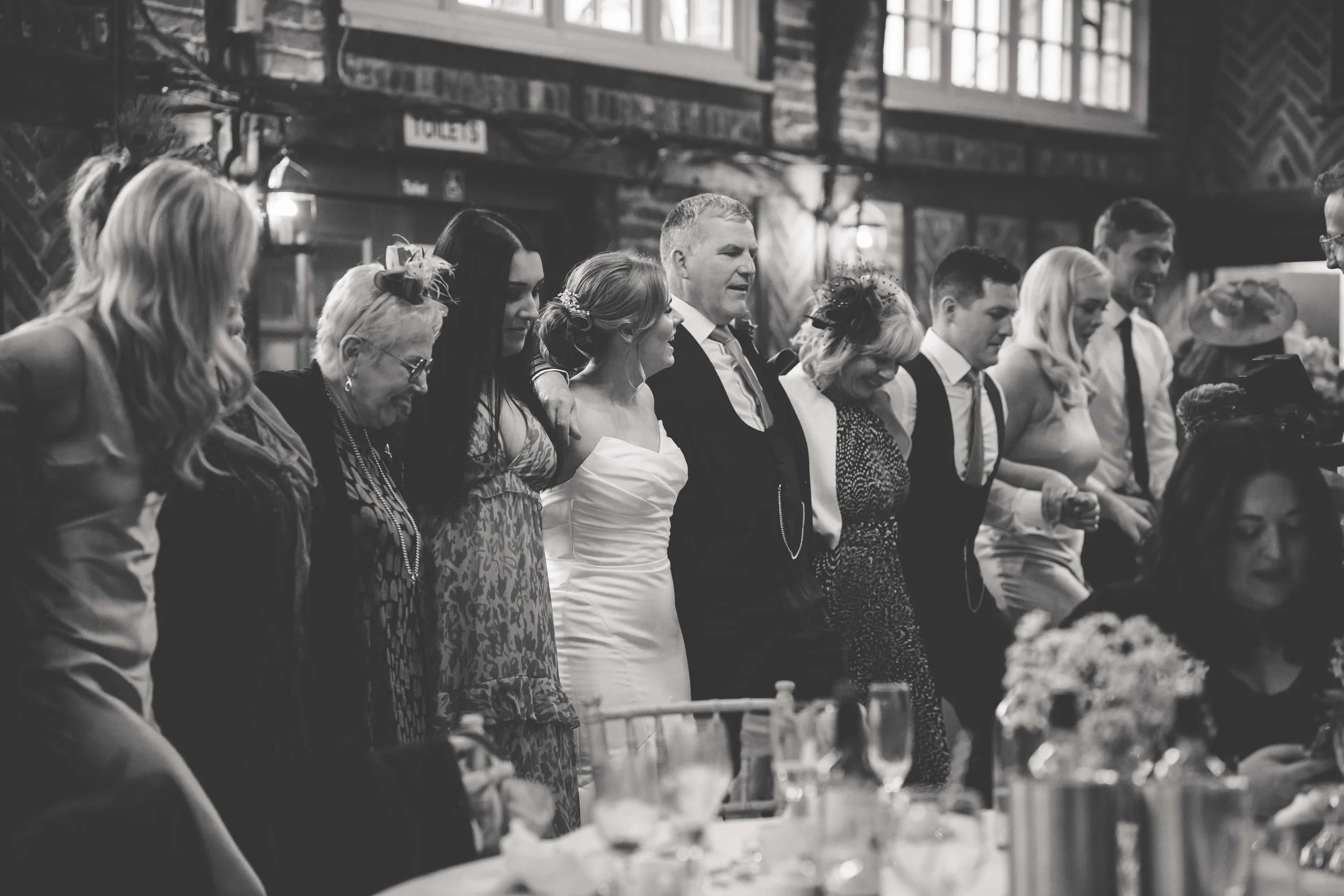 A group of people at a wedding, standing at a reception table, holding each other, and smiling in a rustic indoor setting.