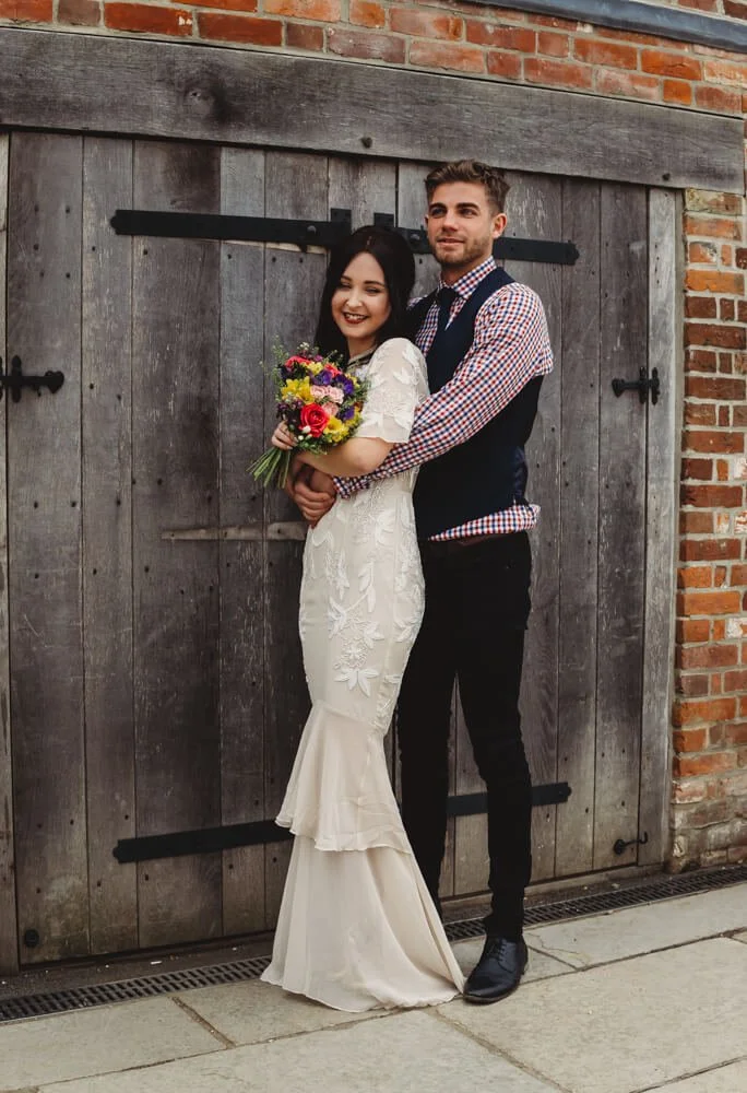 A happy couple standing in front of a rustic wooden barn door; the woman is in a white wedding dress holding a colorful bouquet, and the man is in a checkered shirt with a vest, hugging her from behind.