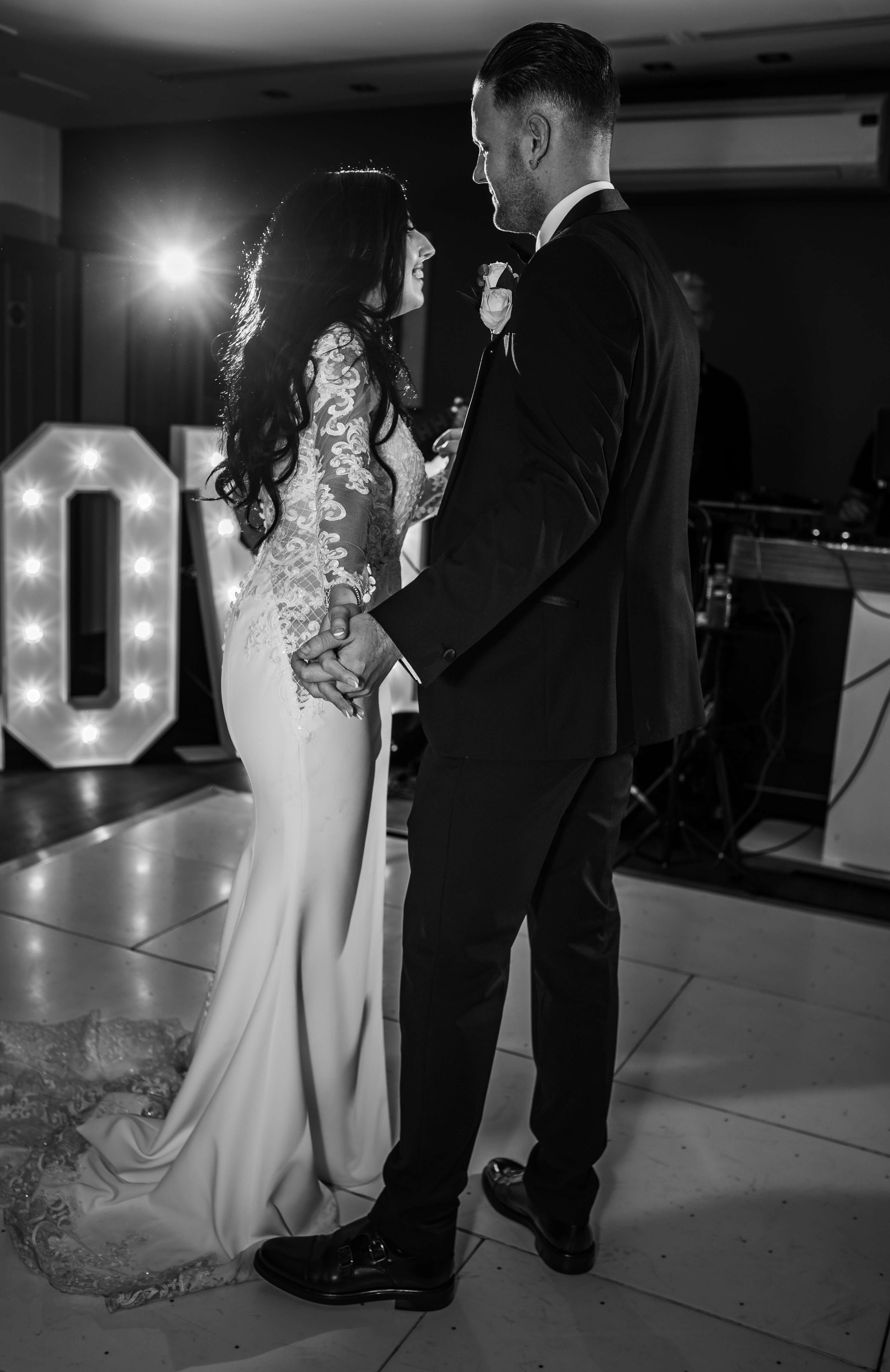 A bride and groom holding hands and dancing at their wedding reception, with large illuminated letters in the background.