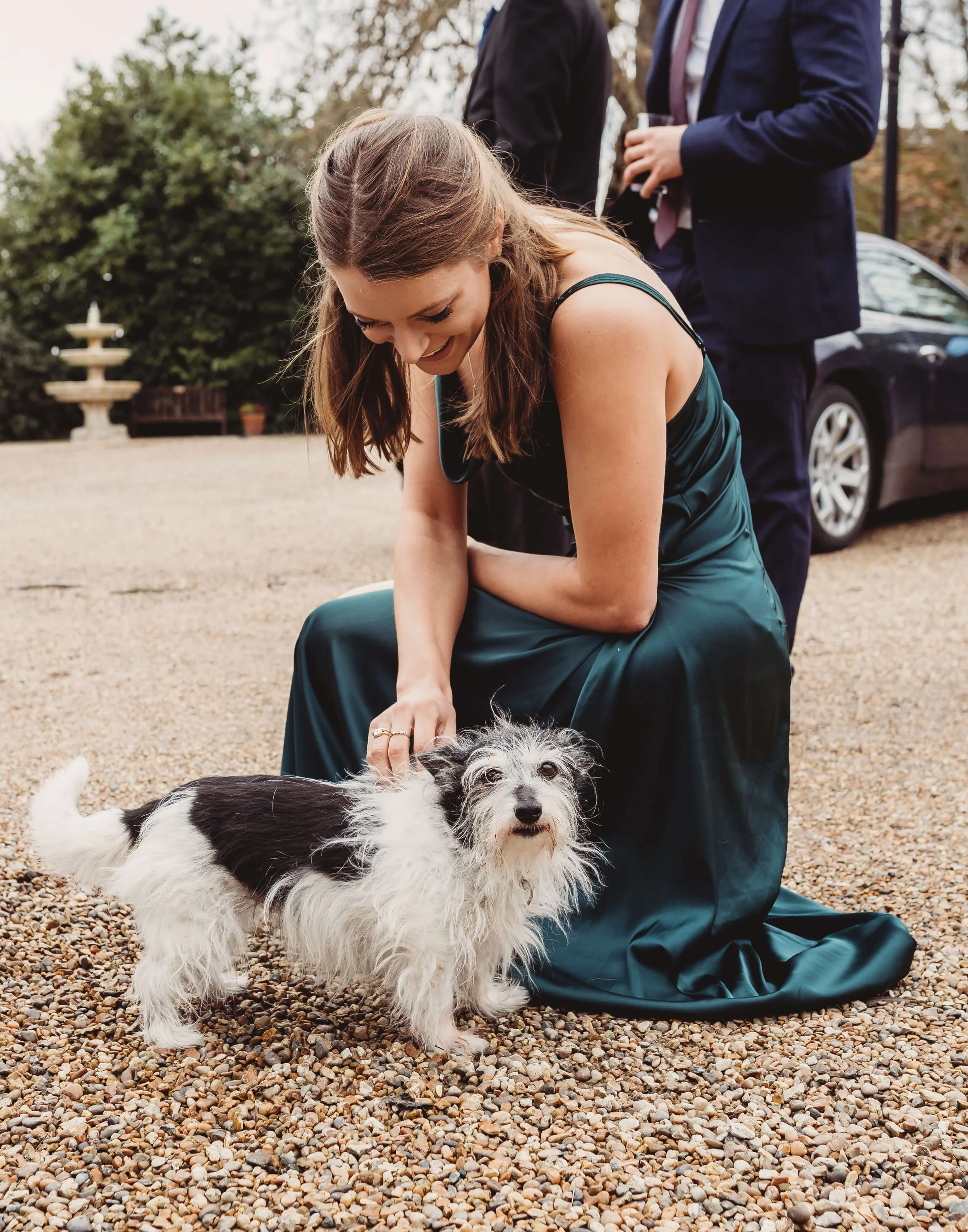 A woman in a teal gown kneels on a gravel surface, petting a black and white dog, with two men in suits standing in the background near a car.