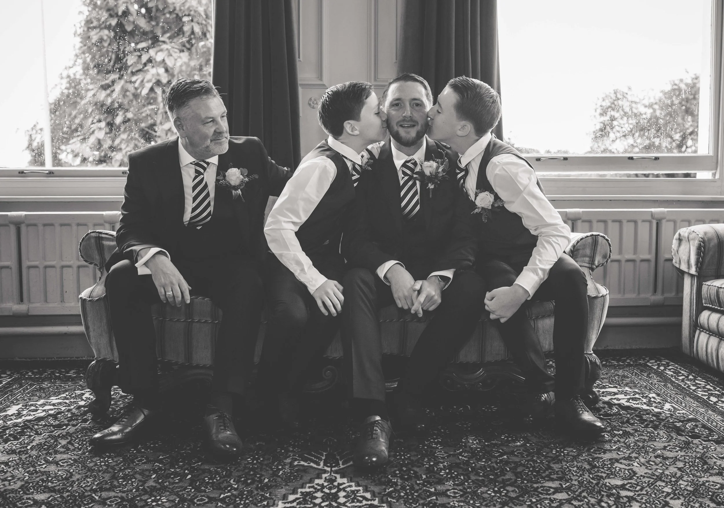A black-and-white photo of five men in suits and ties sitting on a vintage sofa. The central man is being kissed on both cheeks by two young boys, while two other men sit beside him, one looking at the children and the other looking away. The scene i