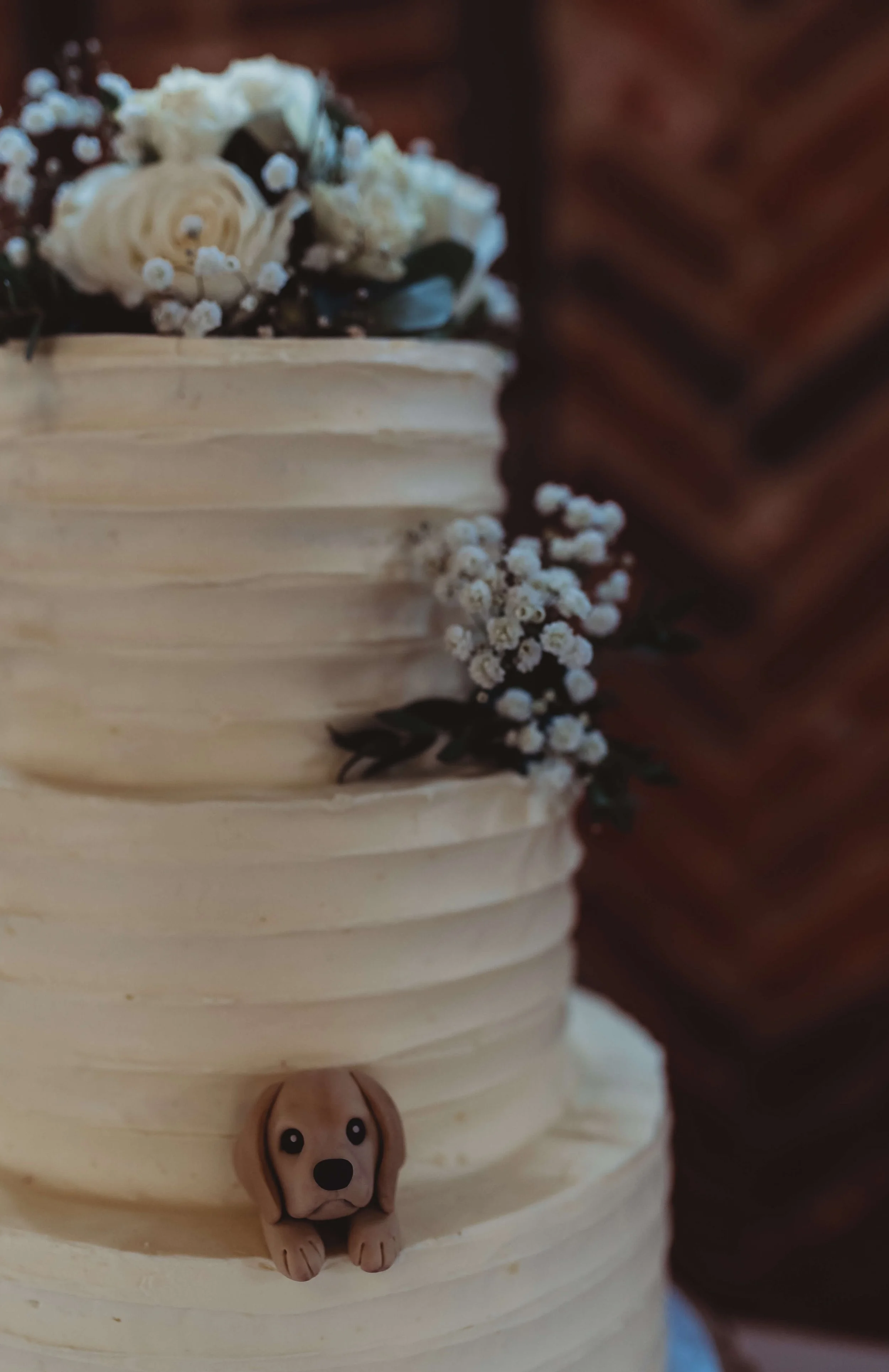 A cream-colored, textured wedding cake decorated with white flowers and greenery. There is a small, cute dog-shaped topper at the bottom of the cake.
