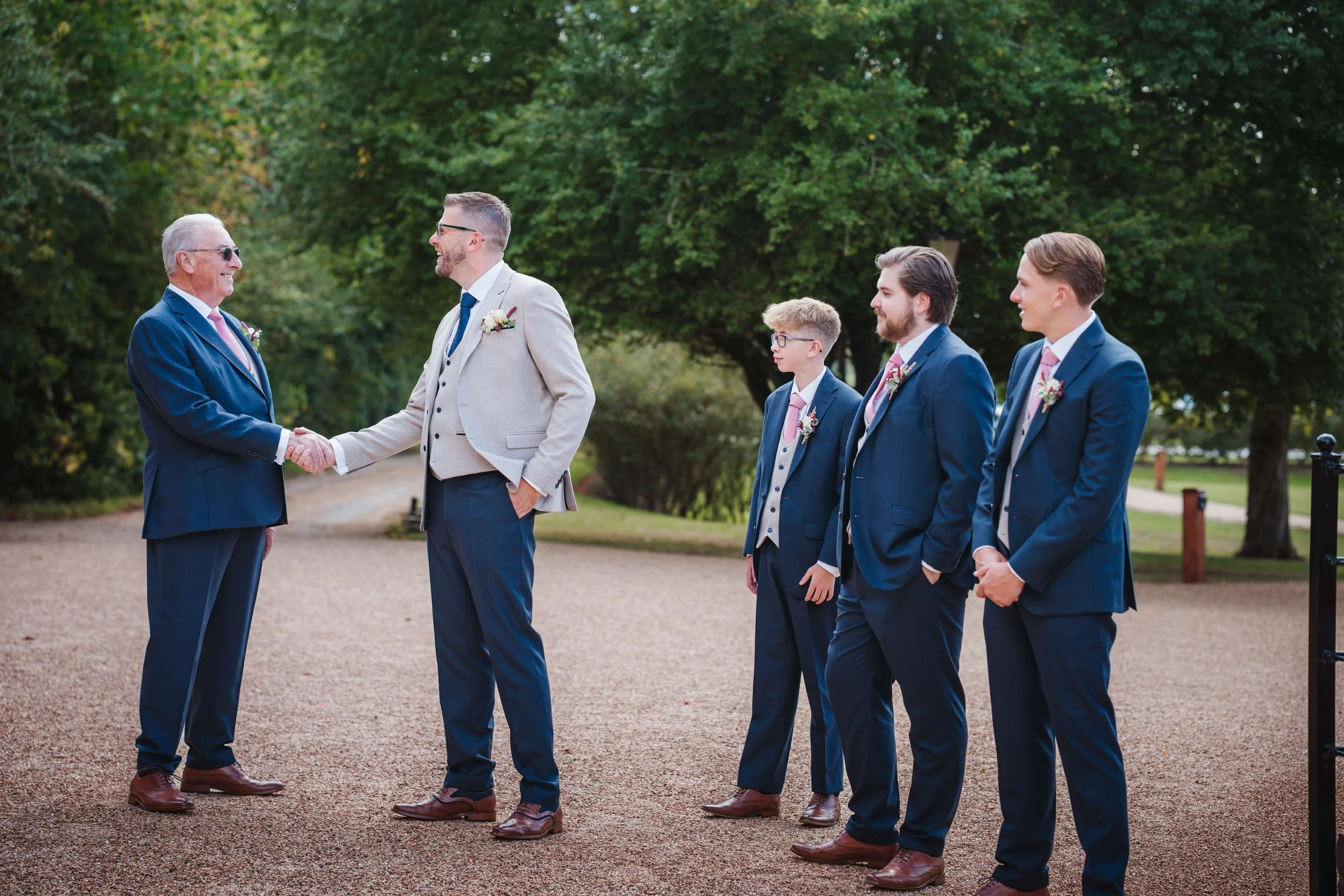 A group of five men dressed in suits, with two older men shaking hands and three younger men standing in line, outdoors in a park with trees in the background.