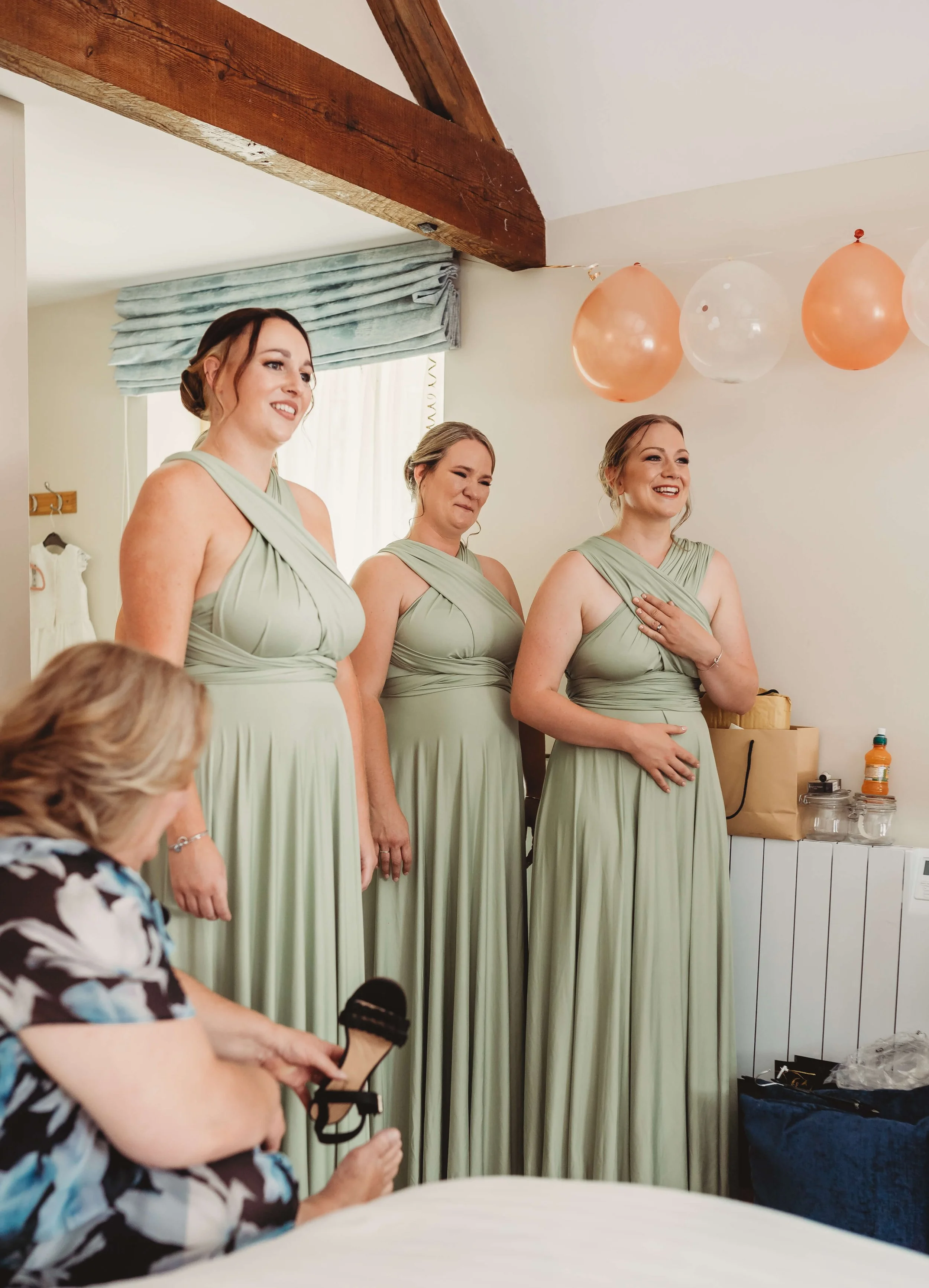 Three women in matching light green dresses are standing side by side, smiling, with a woman in a floral dress sitting in front of them, clipping her microphone. There are pink and clear balloons hanging decorated on the ceiling.