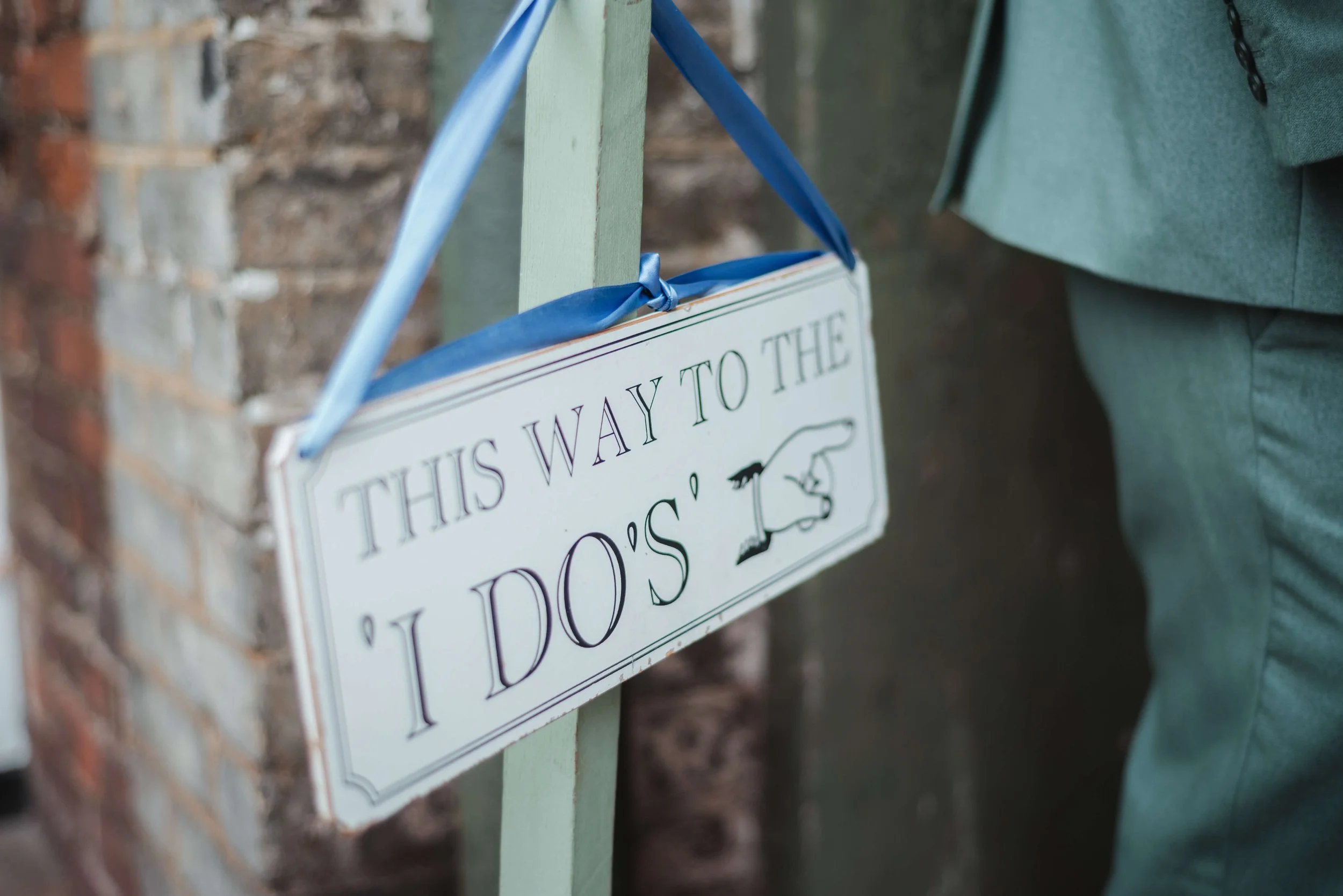 A white rectangular sign hanging with a blue ribbon, pointing to the right, reading 'This way to the D's' and featuring a drawing of a hand pointing in that direction, mounted on a green post next to a brick wall.