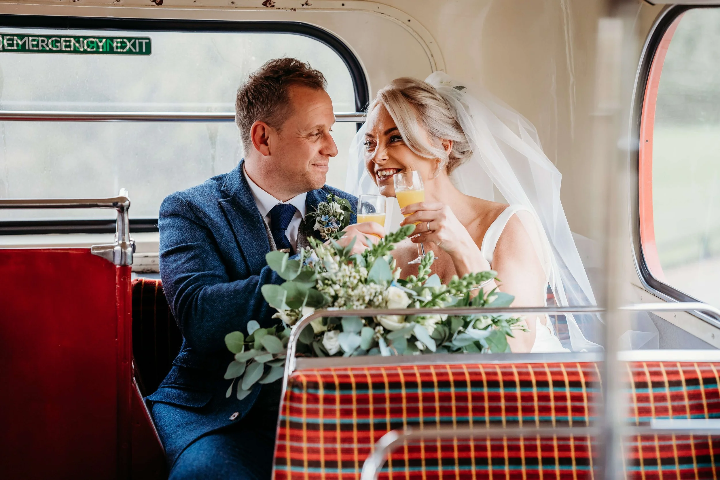 A newlywed couple in wedding attire sharing a toast on a train, with a bouquet of flowers on the table.