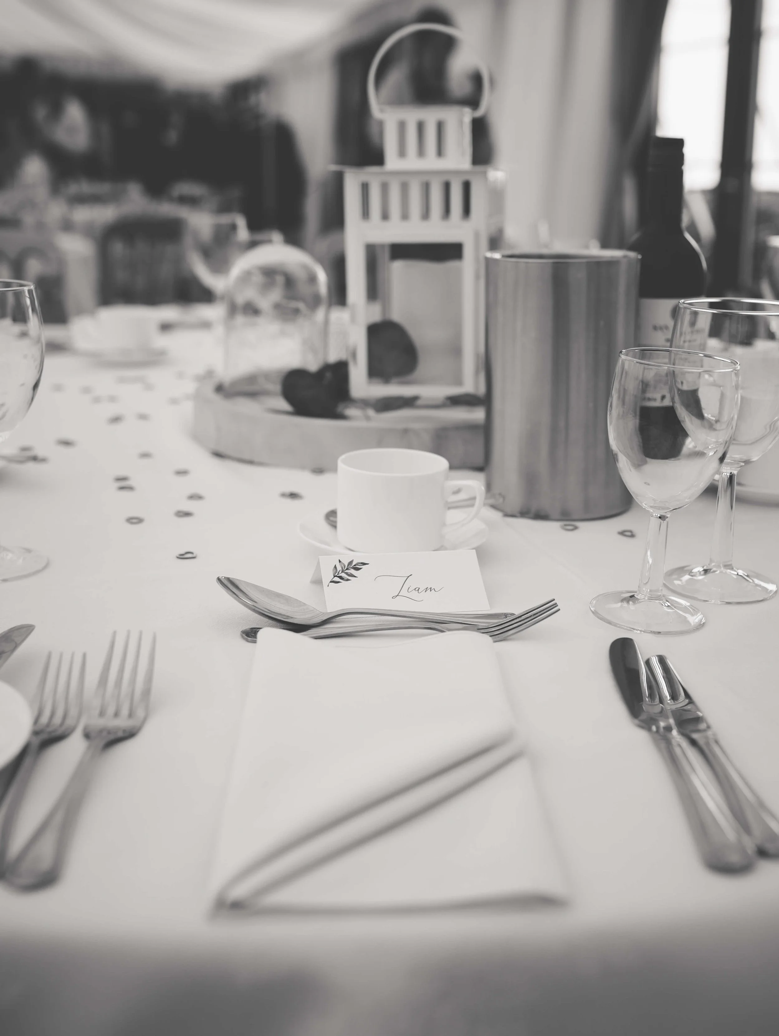 Elegant table setting with silverware, a white napkin, a small card with the name Liam, a white coffee cup, and various glassware. Decor includes a lantern, a bottle, and confetti on a white tablecloth.