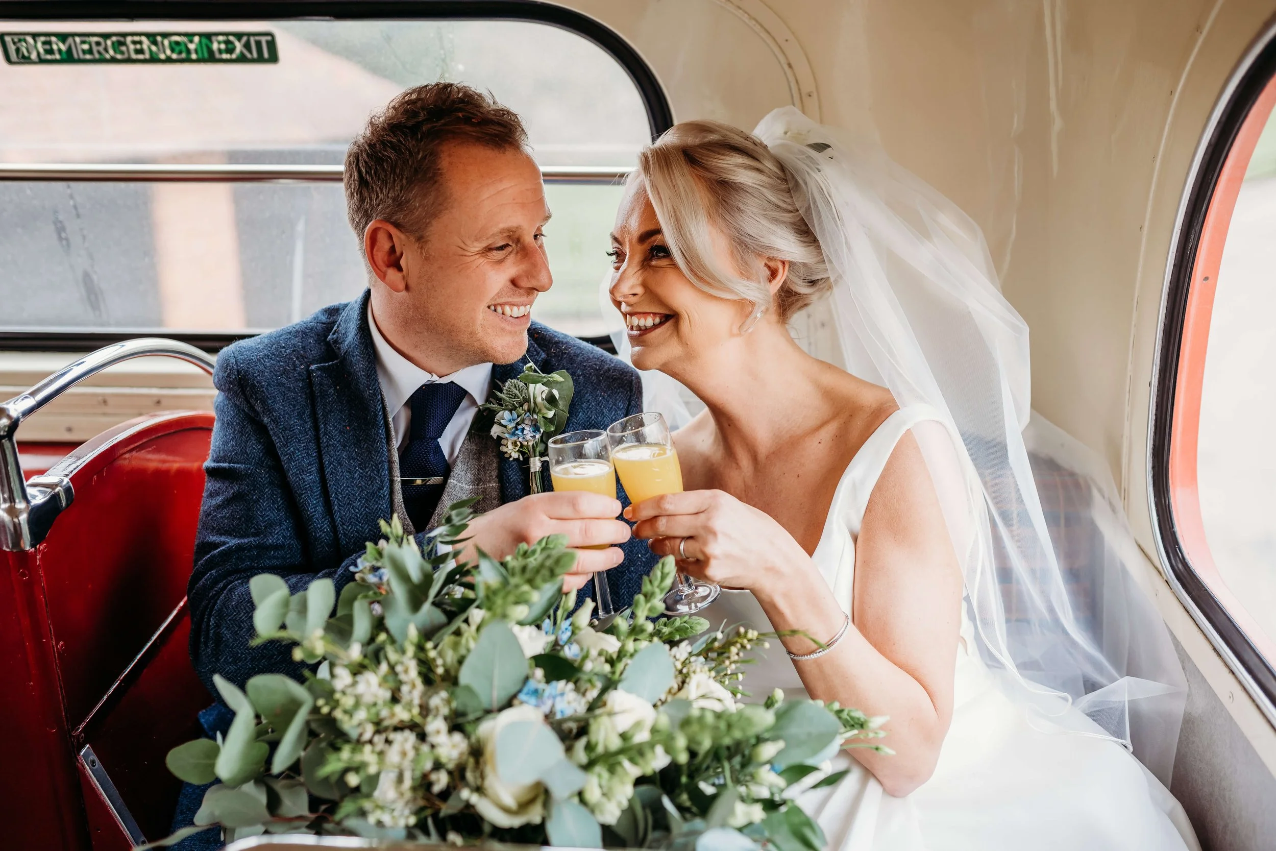Bride and groom celebrating their wedding in a vintage vehicle, clinking glasses of orange juice and smiling at each other.