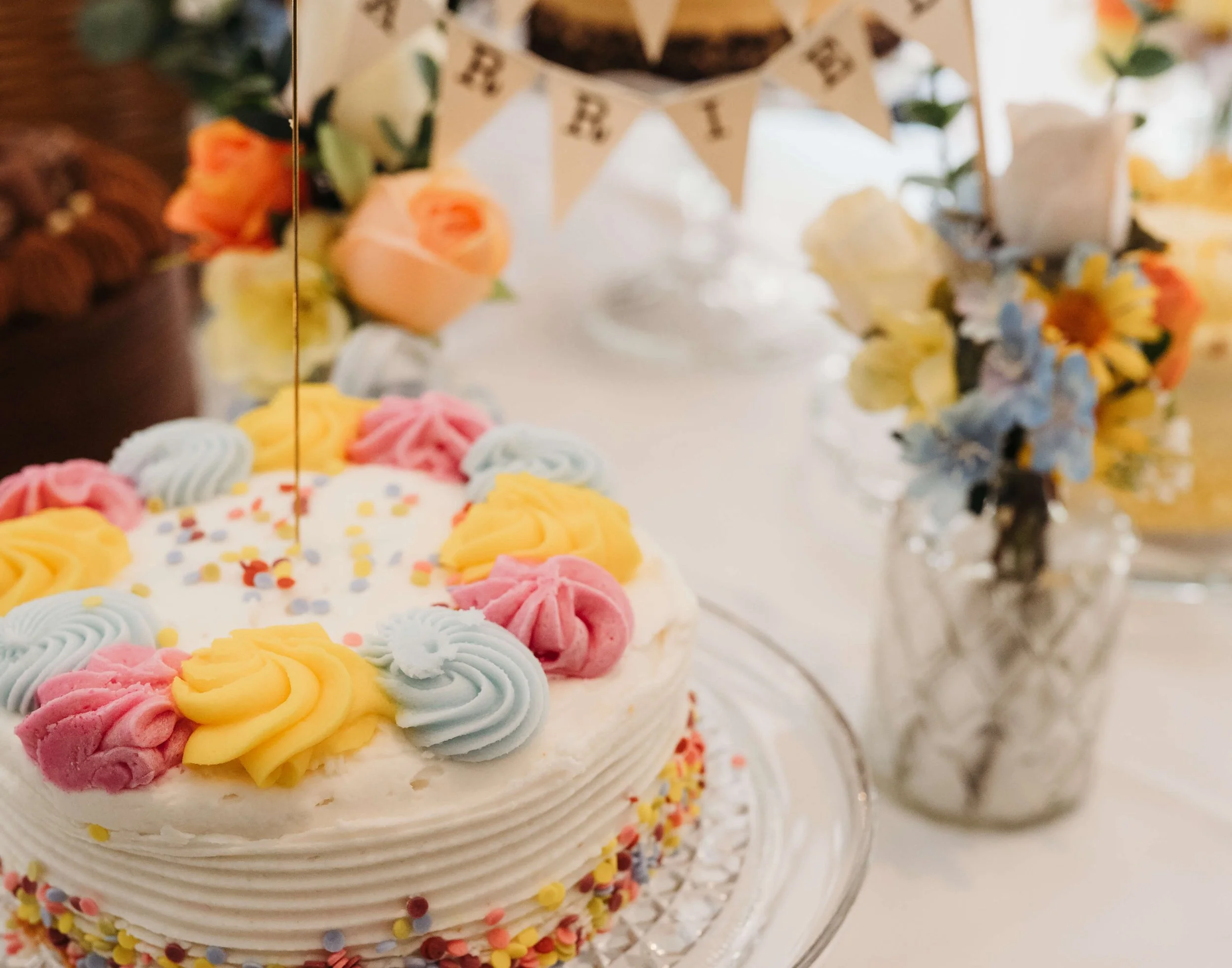 A decorated birthday cake with pastel-colored icing flowers and sprinkles, with a small birthday banner hanging above and floral arrangements in the background.