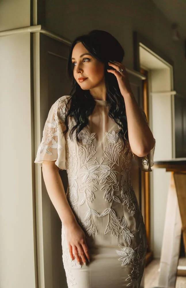 A woman with long black hair, wearing a beige dress with floral embroidery, standing near a window and touching her hair while looking away.