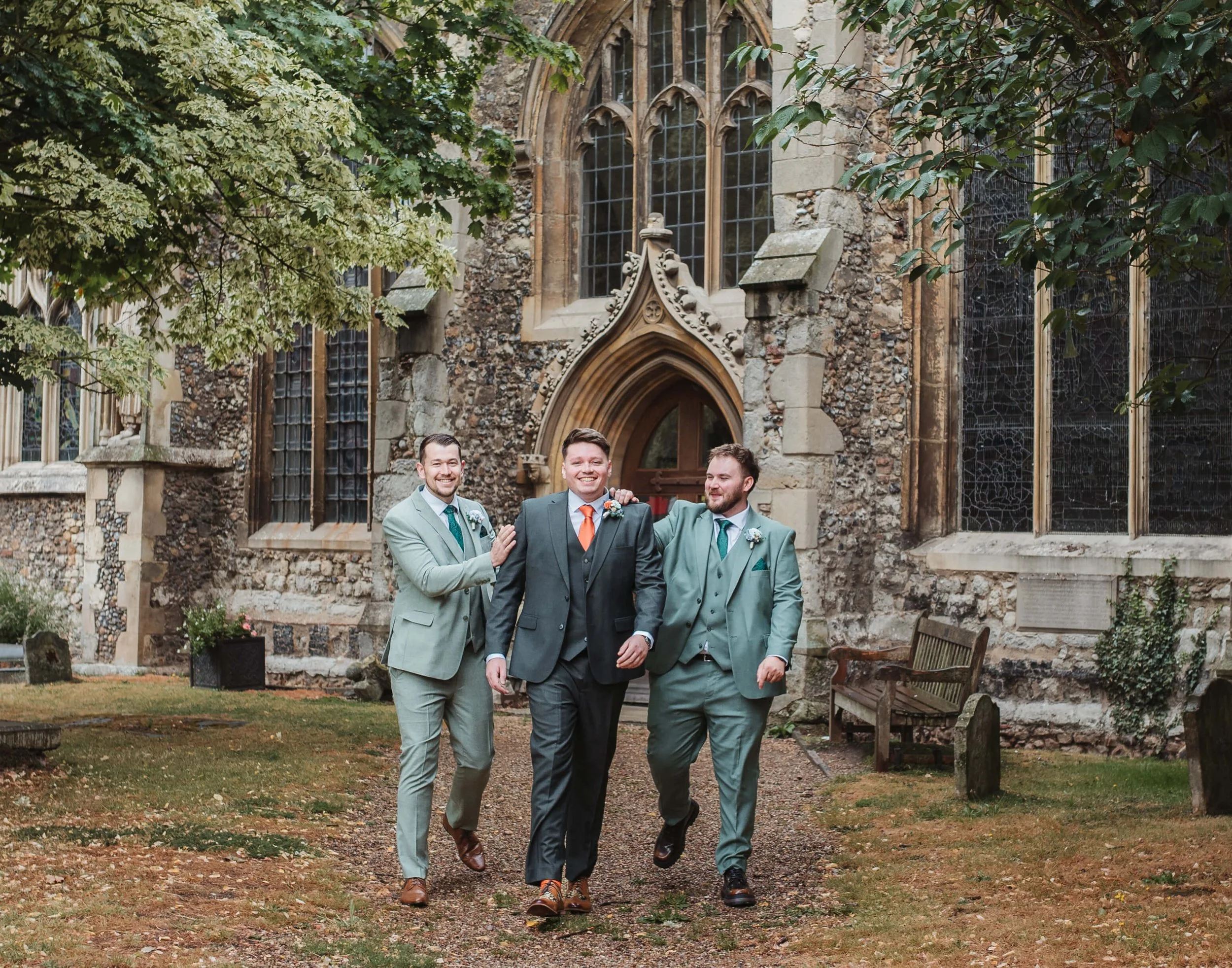 Three men in suits walking and smiling in front of a historic stone church, with green trees and benches nearby.