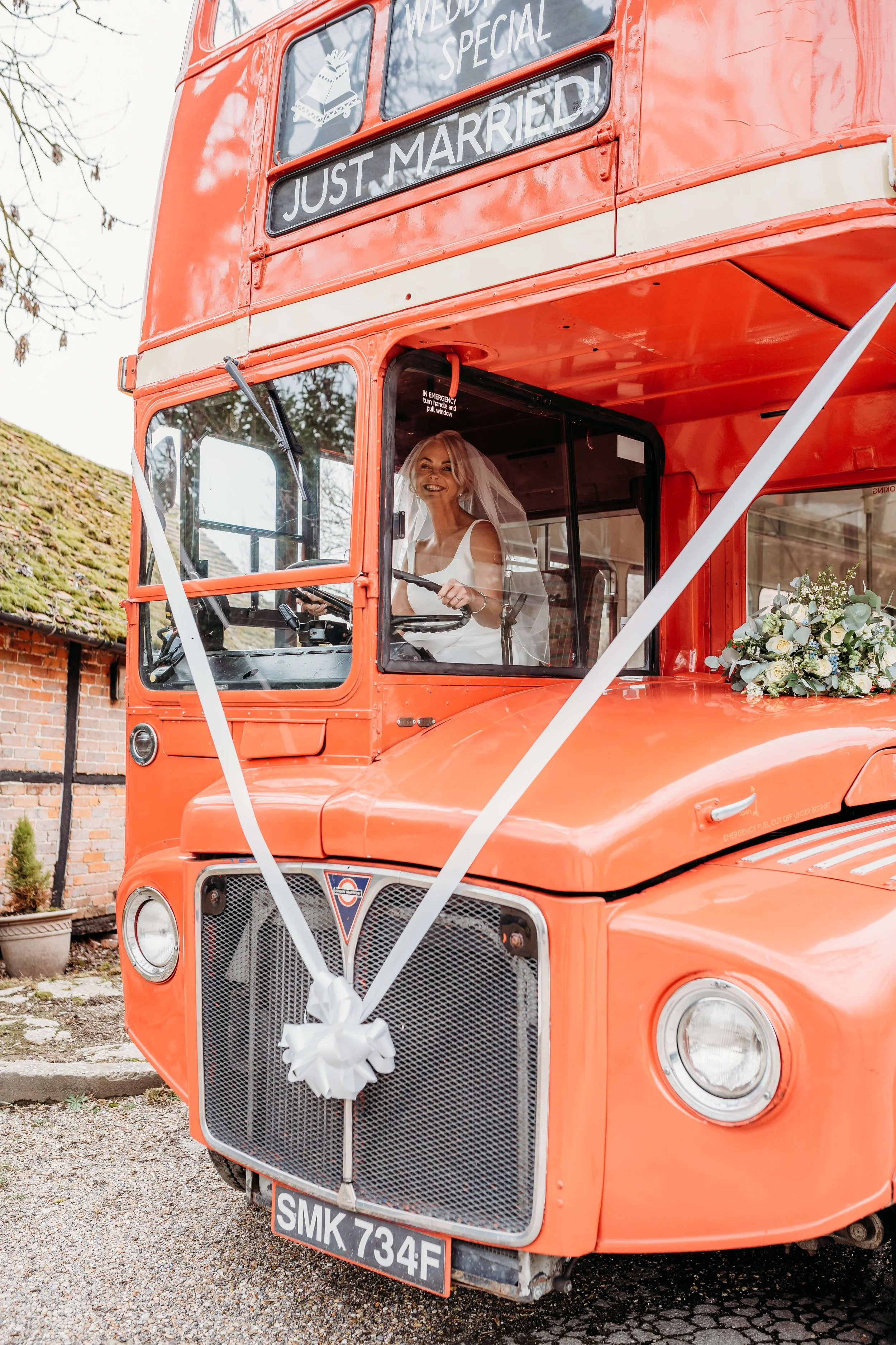 A woman in a wedding dress and veil sitting inside a vintage red double-decker bus decorated with white ribbons and a bouquet of flowers, celebrating her wedding.