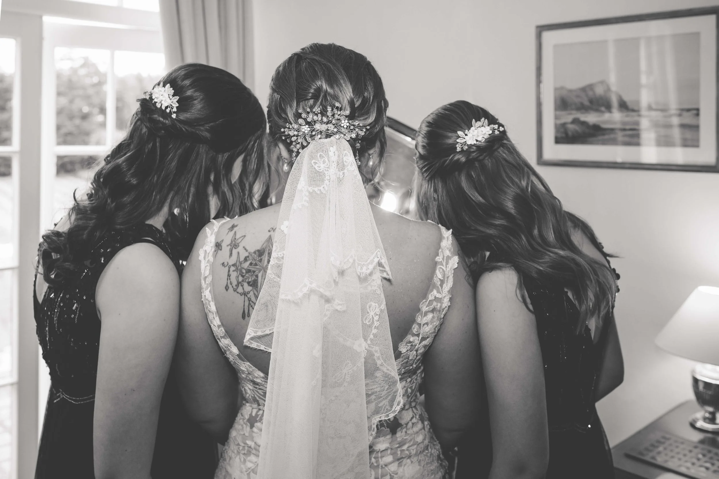 Black and white photo of a bride with three bridesmaids all looking at something on a table in front of them, with a window and framed picture on the wall in the background.