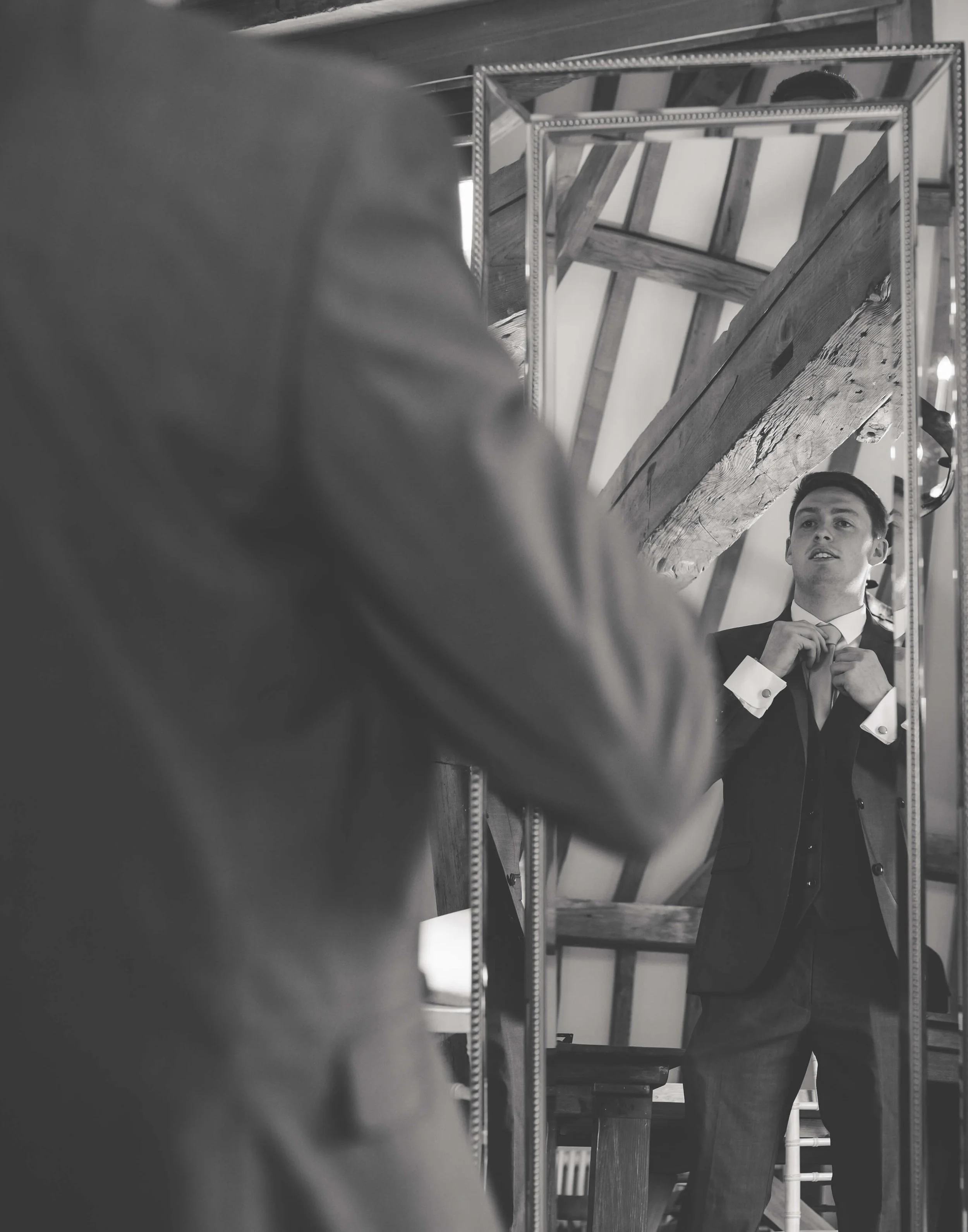 A man in a suit adjusting his tie in front of a mirror, with the reflection showing him. The mirror is surrounded by wooden beams and other mirrors are visible in the background.