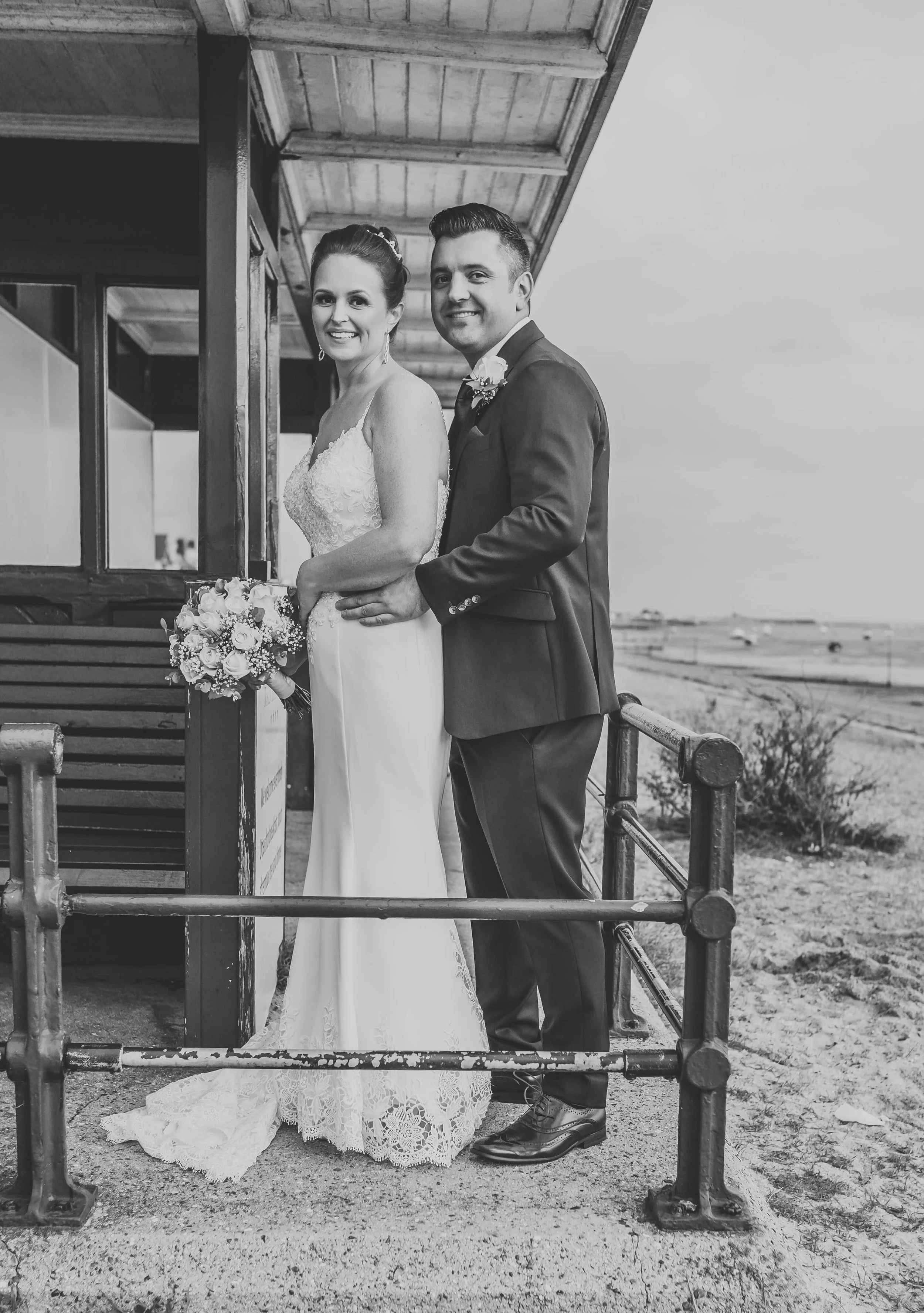 Black and white photo of a bride and groom standing outdoors near a beach, smiling at the camera. The bride is holding a bouquet, wearing a lace wedding dress. The groom is in a suit, standing behind her with his hand on her waist.
