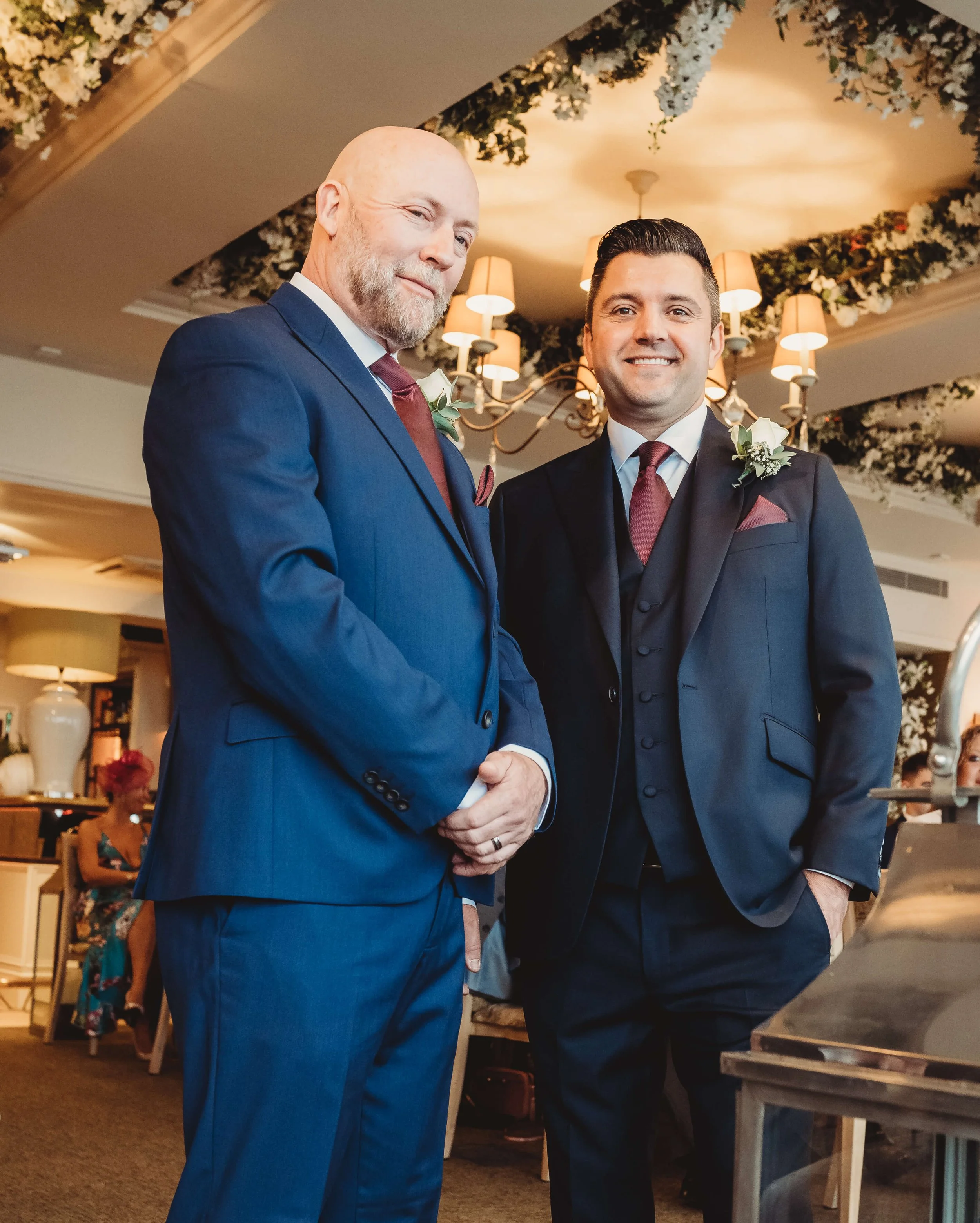 Two men in suits standing side by side at a formal event, smiling at the camera. They are dressed in dark suits with maroon ties and white shirts, each with a boutonniere on his lapel. The background features elegant decor and warm lighting.