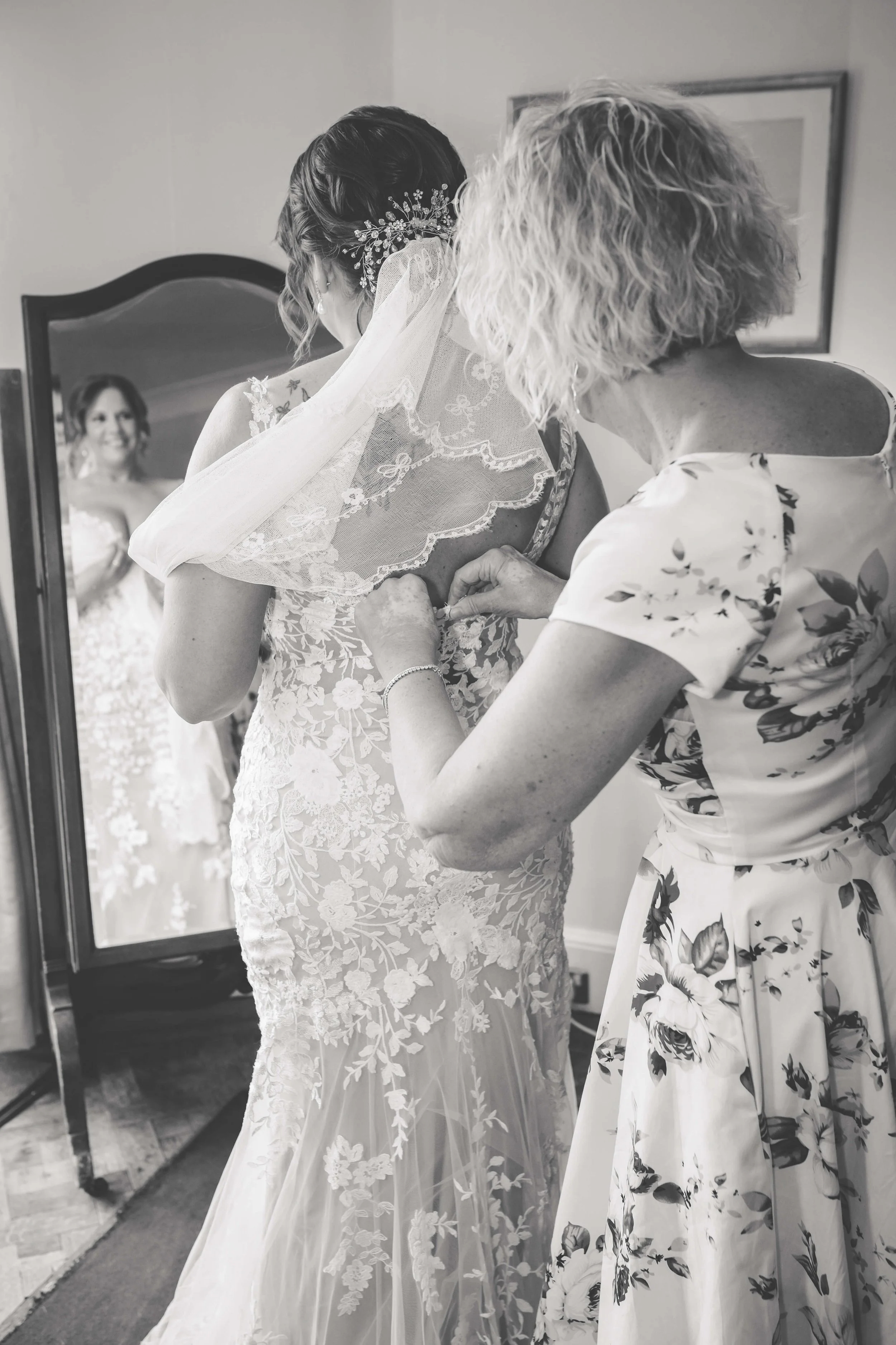 A woman, possibly a bride, is getting ready with help from another woman, possibly her mother, as she adjusts her wedding dress. The woman is looking at herself in a mirror.