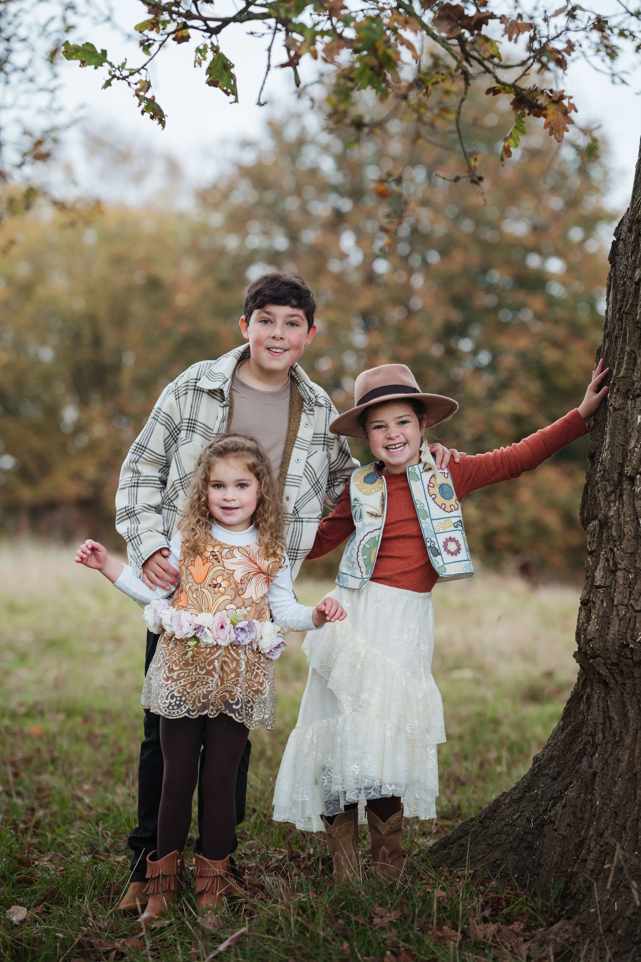 A family of three children standing outdoors in a park with autumn foliage, smiling and posing near a tree.